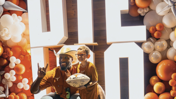 A man in a cowboy hat poses with a child holding a white, signed football. The pair is in front of big, light-box letters that read "DTE 100," surrounded by a burnt-orange and white balloon arch.