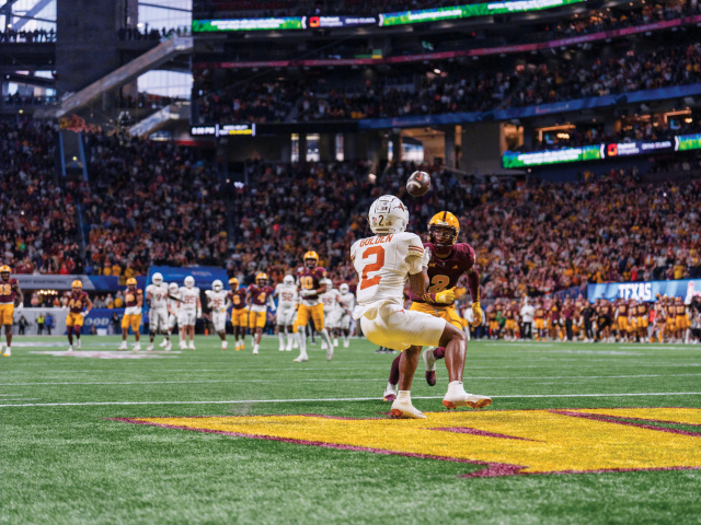 A Longhorn football player in white catches a football ahead of a football player in maroon.