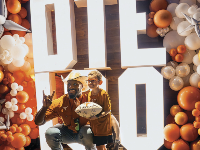 A man in a cowboy hat poses with a child holding a white, signed football. The pair is in front of big, light-box letters that read "DTE 100," surrounded by a burnt-orange and white balloon arch.