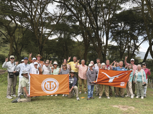 A group of elderly travelers pose in front of a wooded background, holding flags with the Texas Exes logo and Longhorn, respectively.