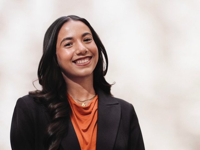 A young woman in a burnt-orange, cowl-necked blouse under a black blazer smiles against a dappled, white background.