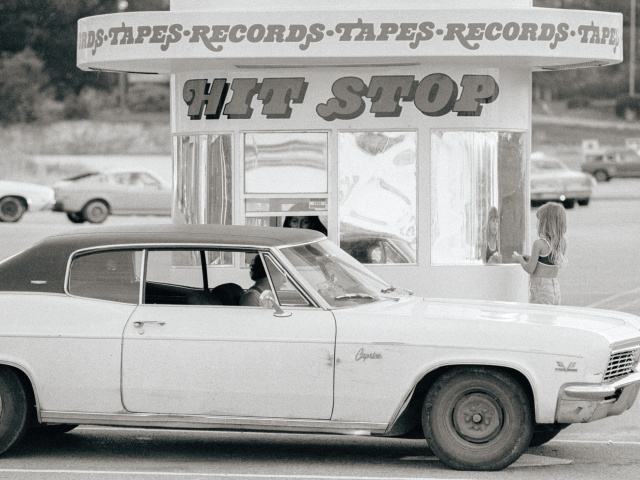 A black and white image of an old car pulled up in front of a drive-through record store with a sign that says HIT STOP in a groovy font.