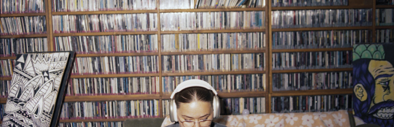 A young woman wearing overhead earphones on a couch, in front of a full wall of shelves packed with CDs.