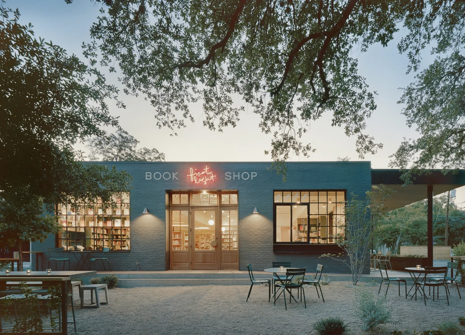 Viewed through the branches of oak trees, warm light shines from inside a one-story concrete building with a red neon script sign that says "First Light Books".