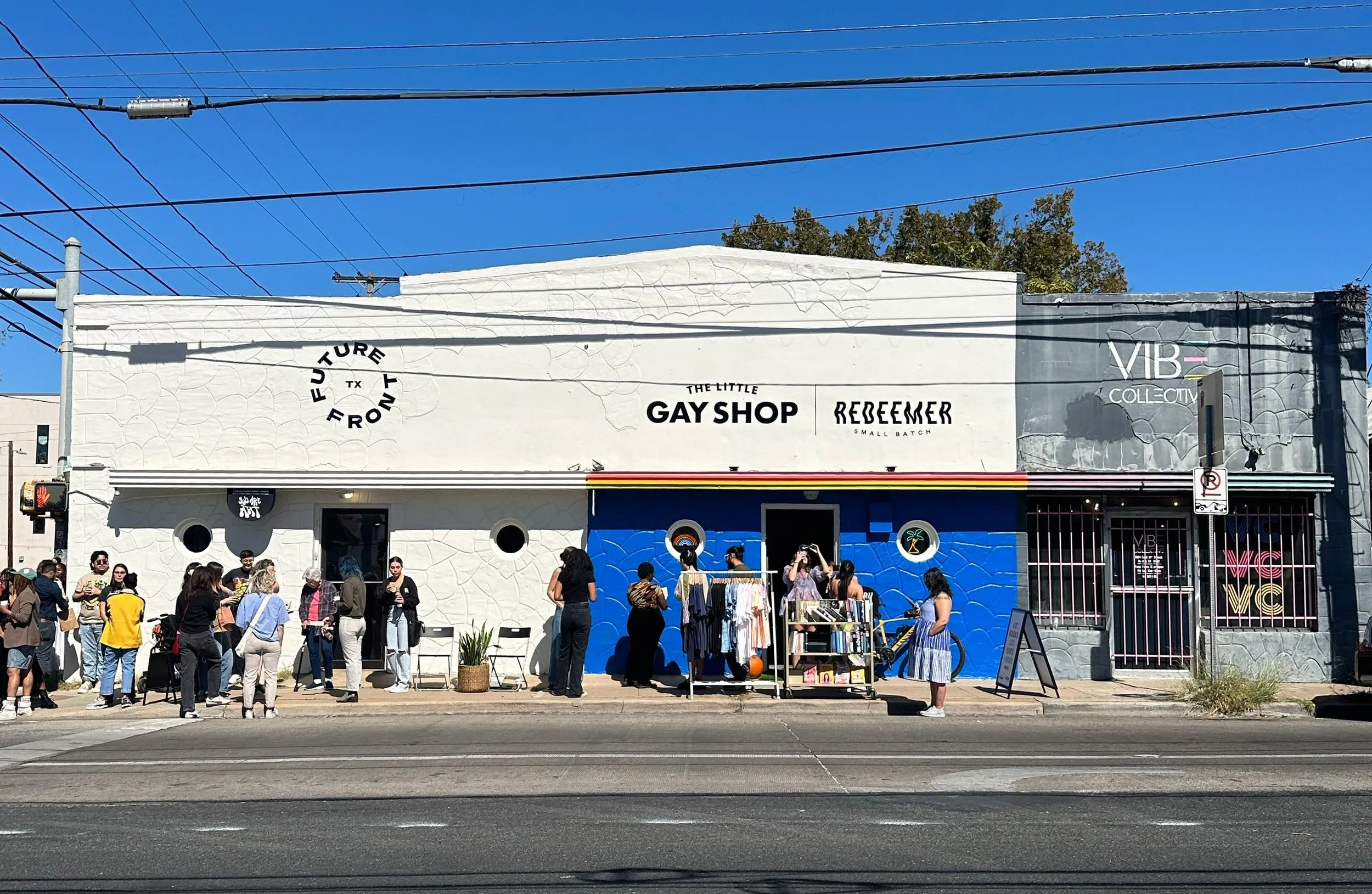People gather outside of a white stucco storefront that reads "The Little Gay Shop".