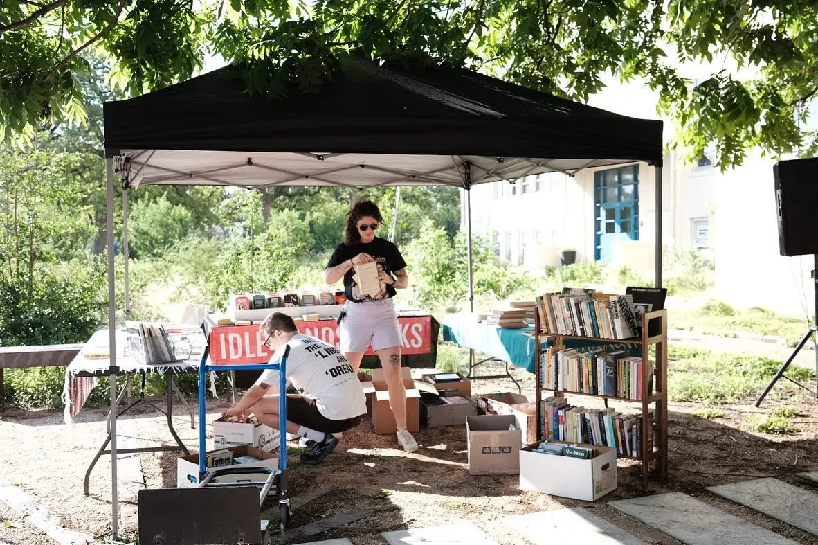 Under tree cover and a black pop-up tent, a woman stands and a man squats in front of short shelves and crates of books.