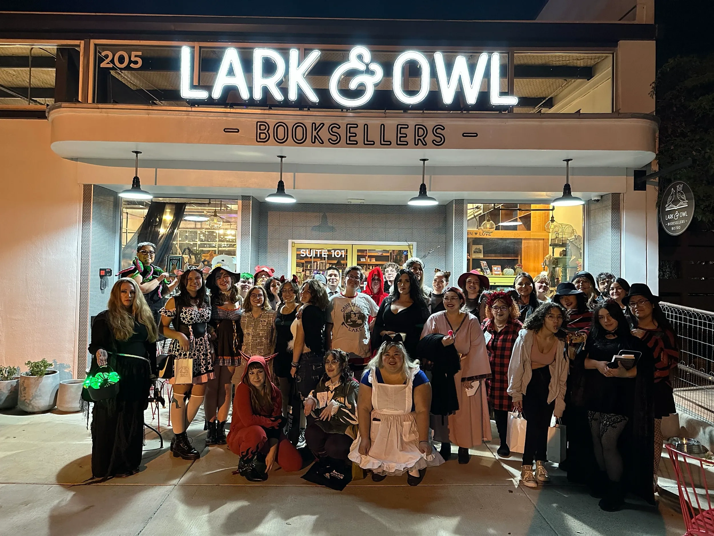 A grou pof people stand in front of a white storefront with a white neon sign reading "Lark & Owl Booksellers".