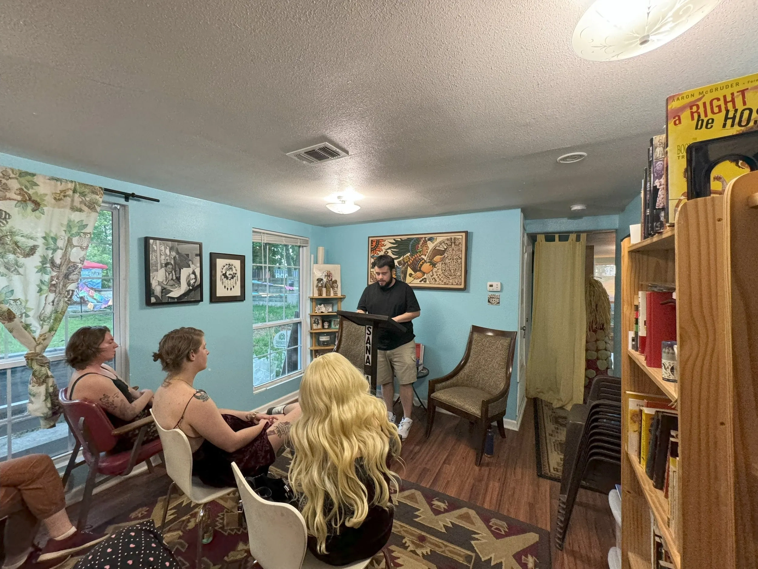 Three women listen to a man reading from a lectern, in a blue room with bookshelves lining one wall and a window on the other.