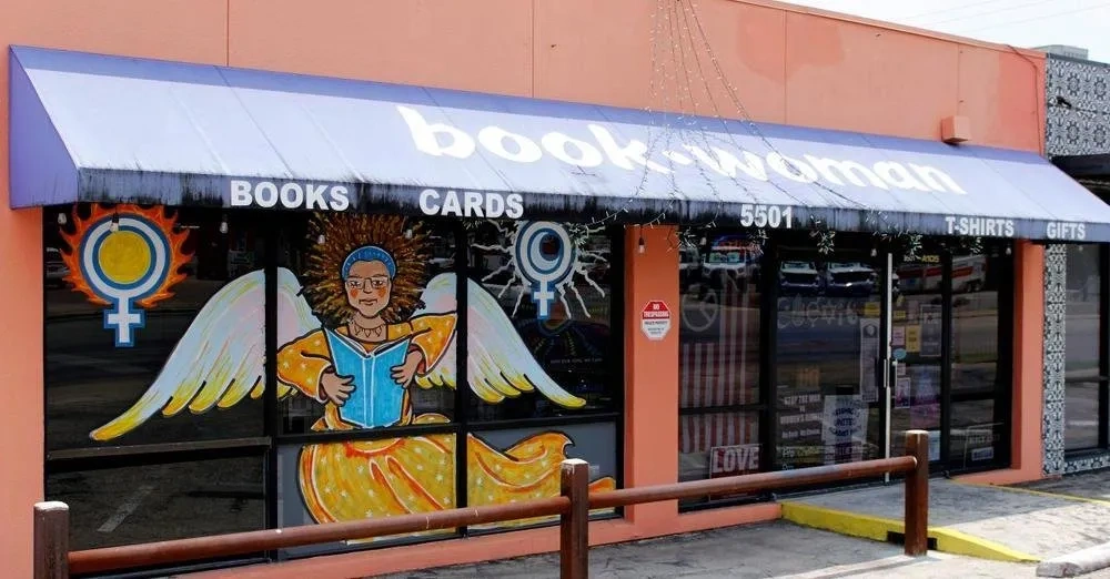 A blue awning reads "book woman" over a glass storefront painted with an angel in a yellow dress reading a book, flanked by symbols for the female sex.