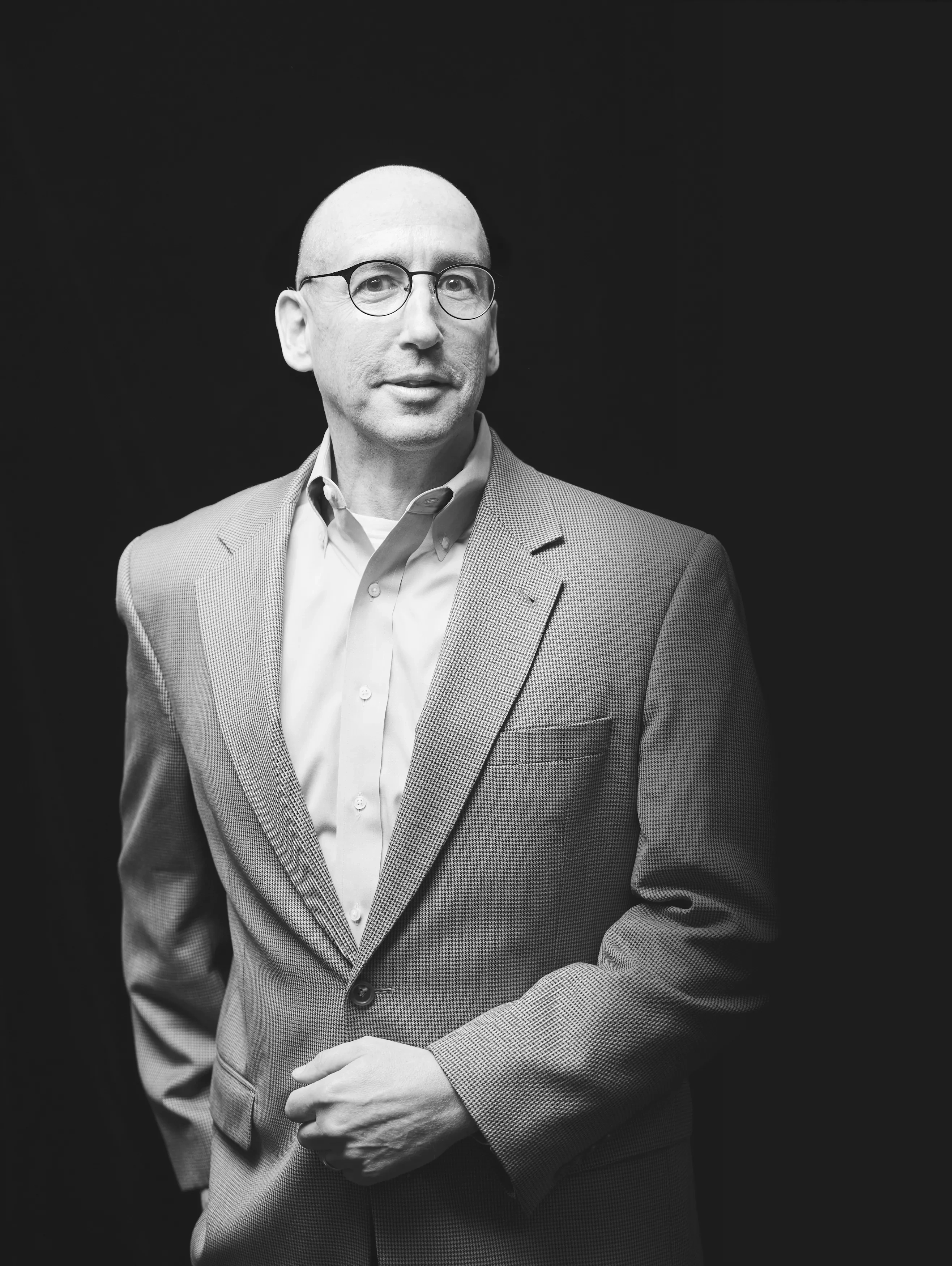 A man in a blazer stands against a black background.