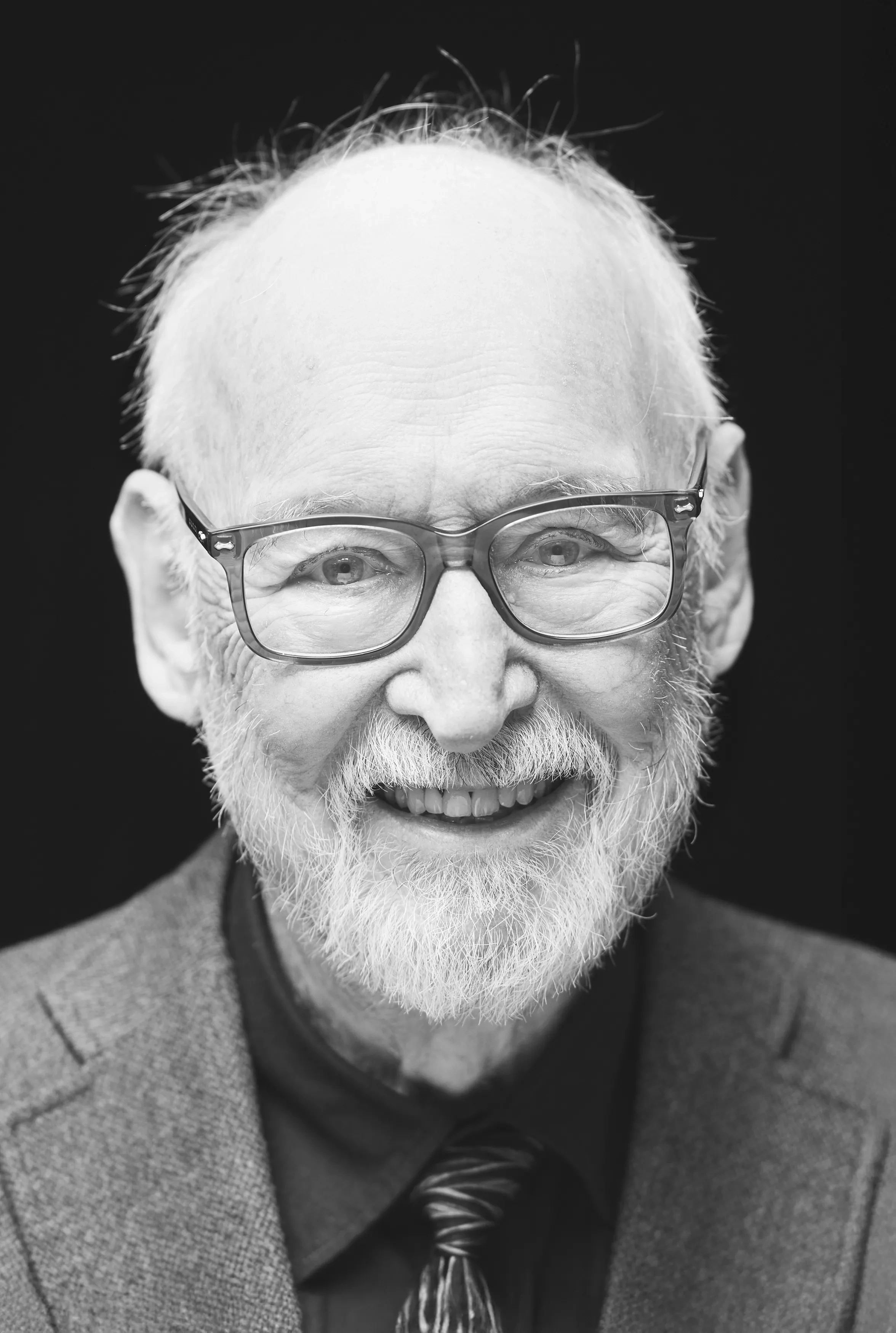 A close-up shot of an old, bearded man wearing glasses against a black backdrop.