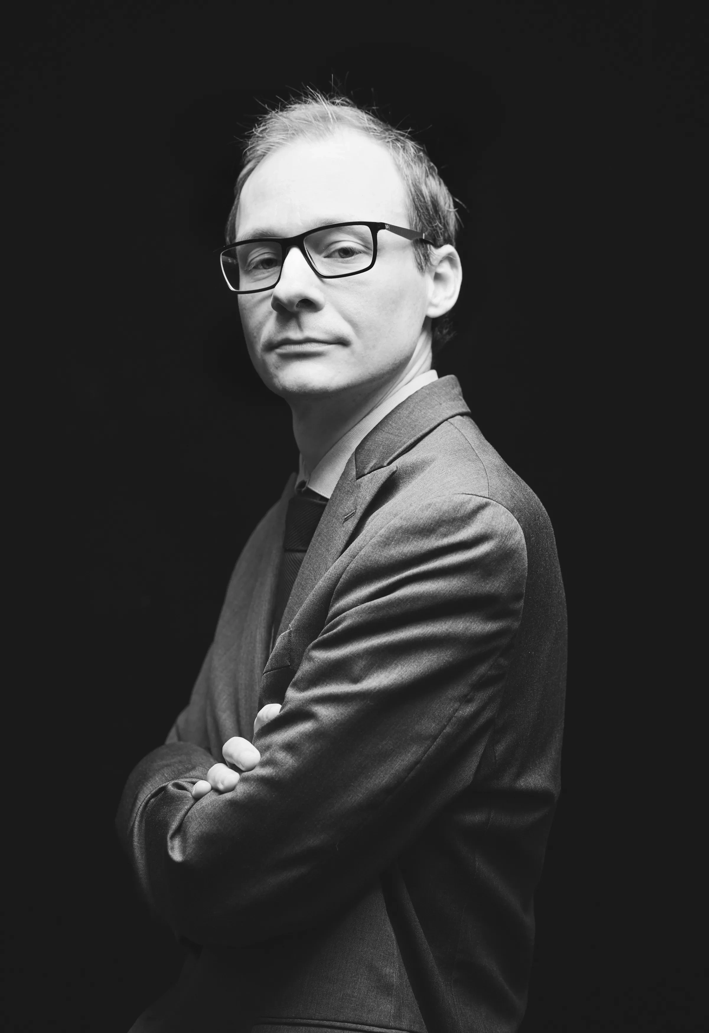 A middle-aged man in square glasses stands in profile, with his head turned toward the camera, unsmiling.