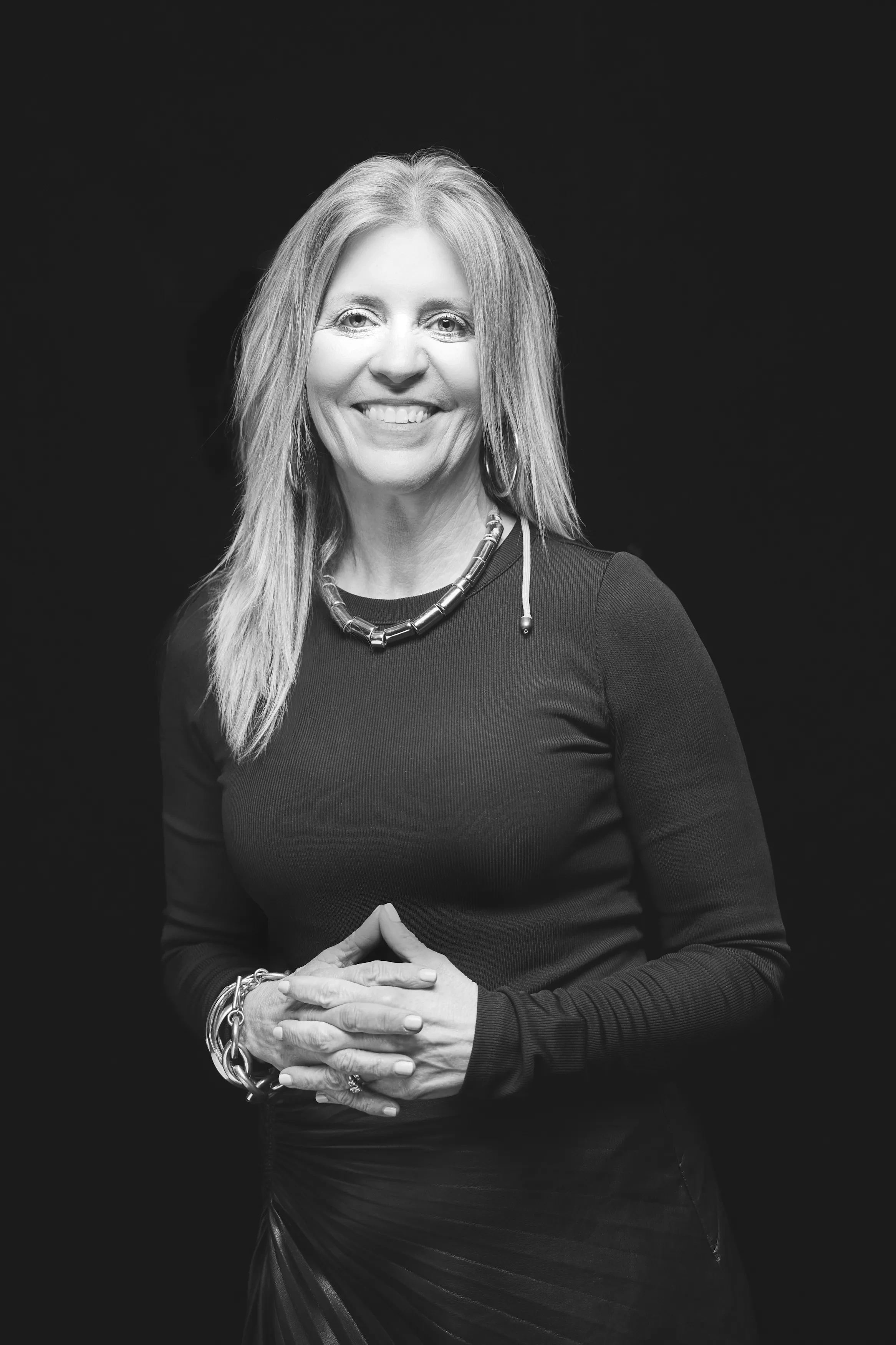 A blonde woman stands with her hands clasped against a black backdrop.