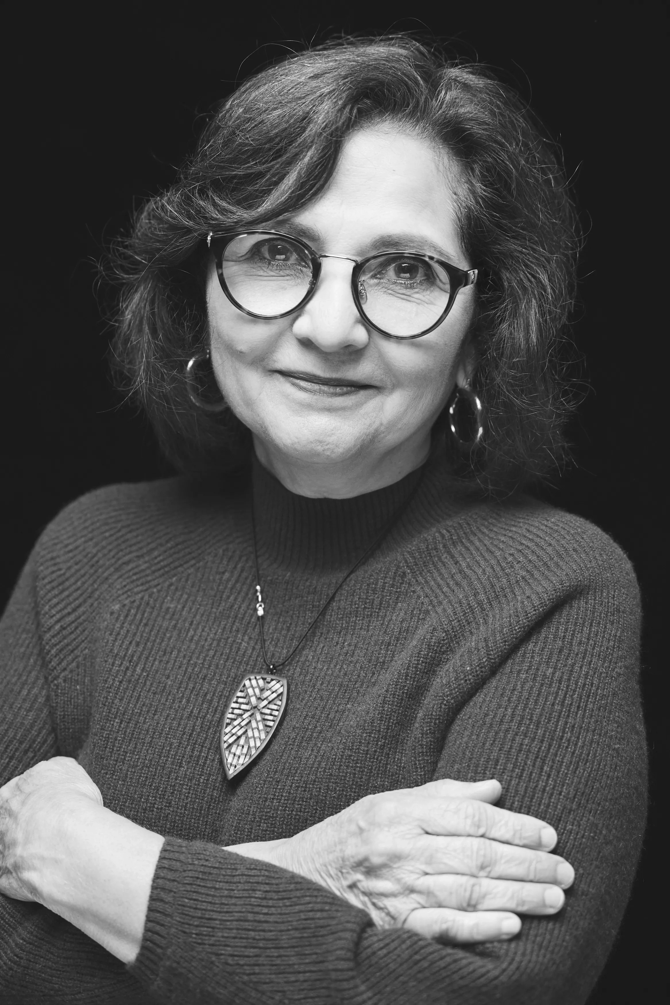 A woman with dark wavy hair in a sweater with a bold necklace stands with her arms around herself in front of a black backdrop.