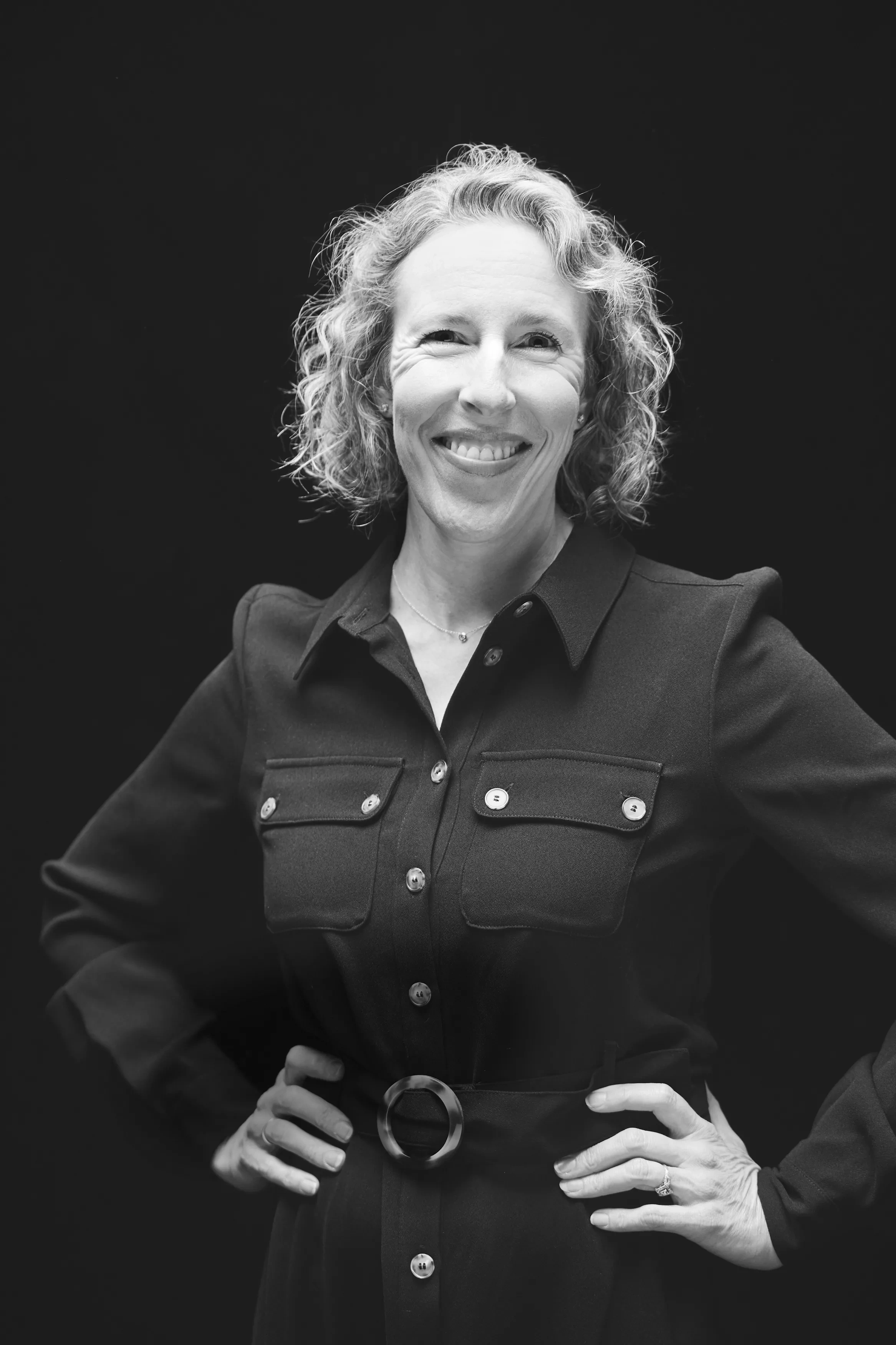 A woman with blonde curly hair stands against a black backdrop with her hands on her hips.