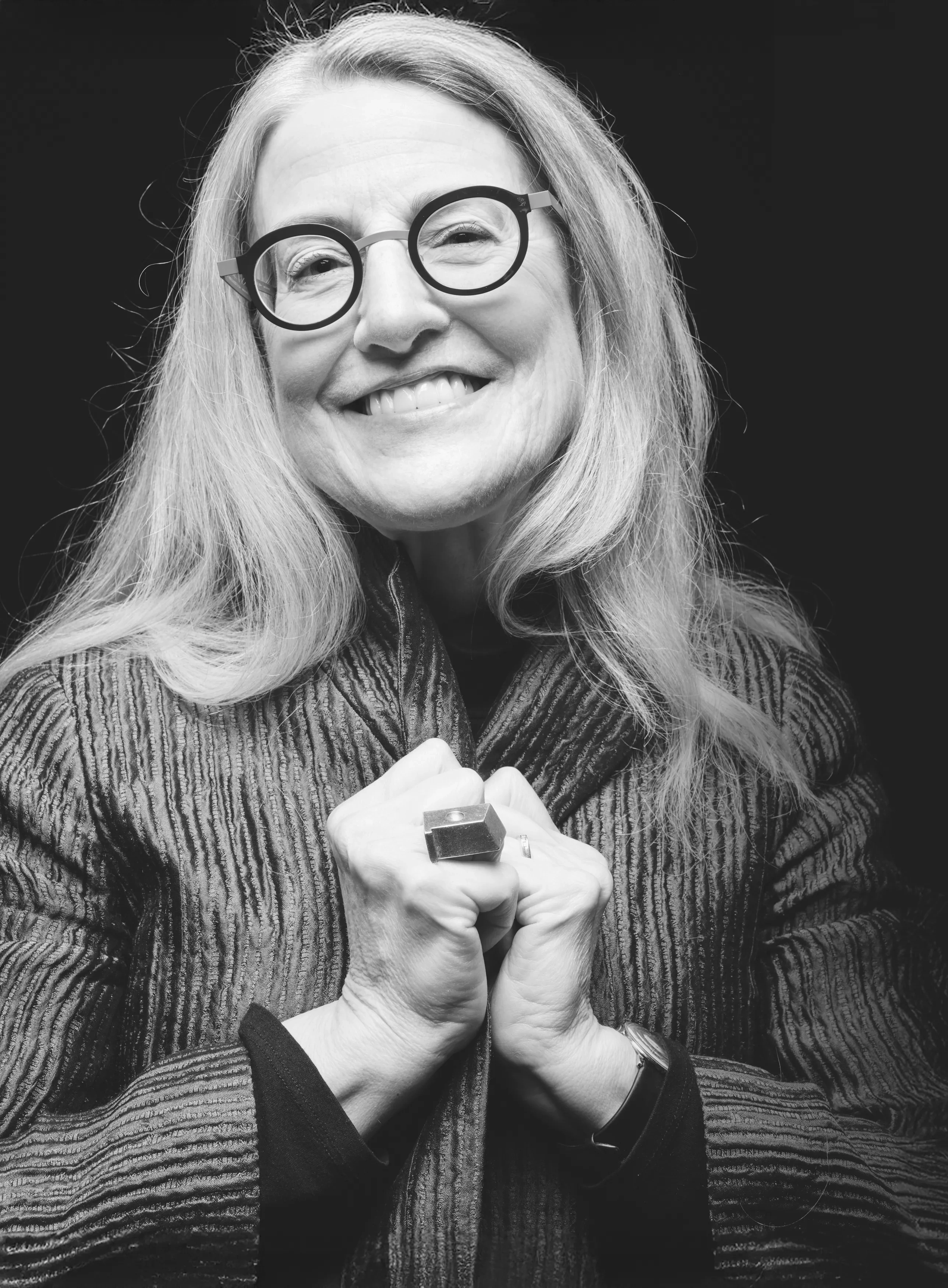 A smiling woman with shoulder-length gray hair and bold black and gold glasses stands with her hands clasped at her clavicle against a black backdrop.