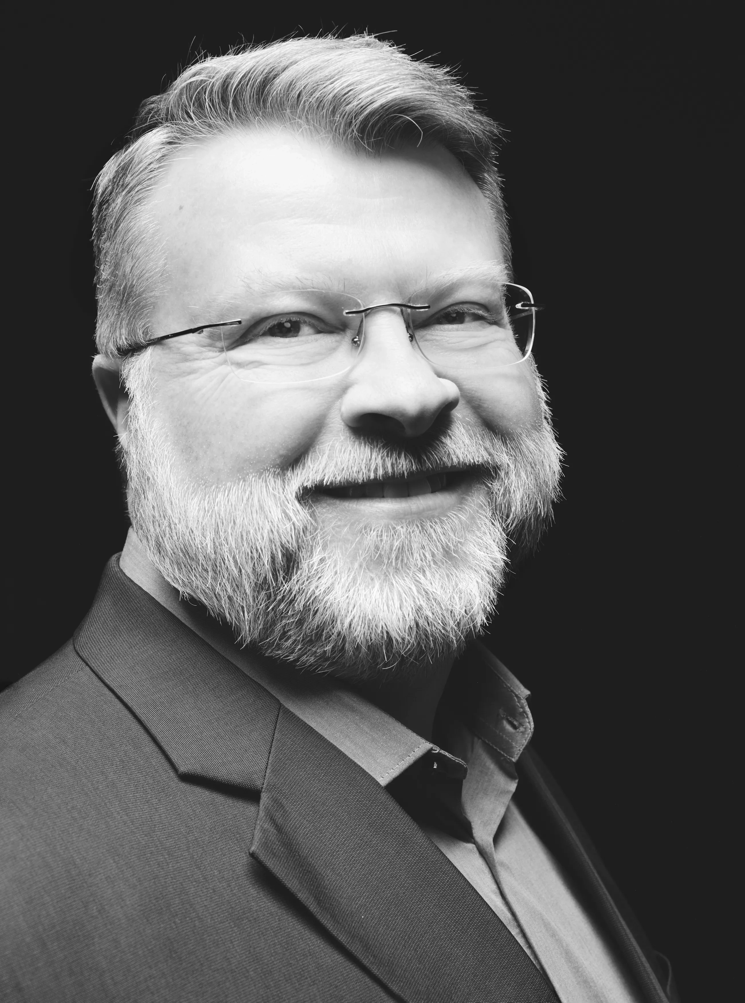 A close-up shot of a middle-aged man in rimless glasses with a tight beard, standing diagonally toward the camera in a sport coat and button down.