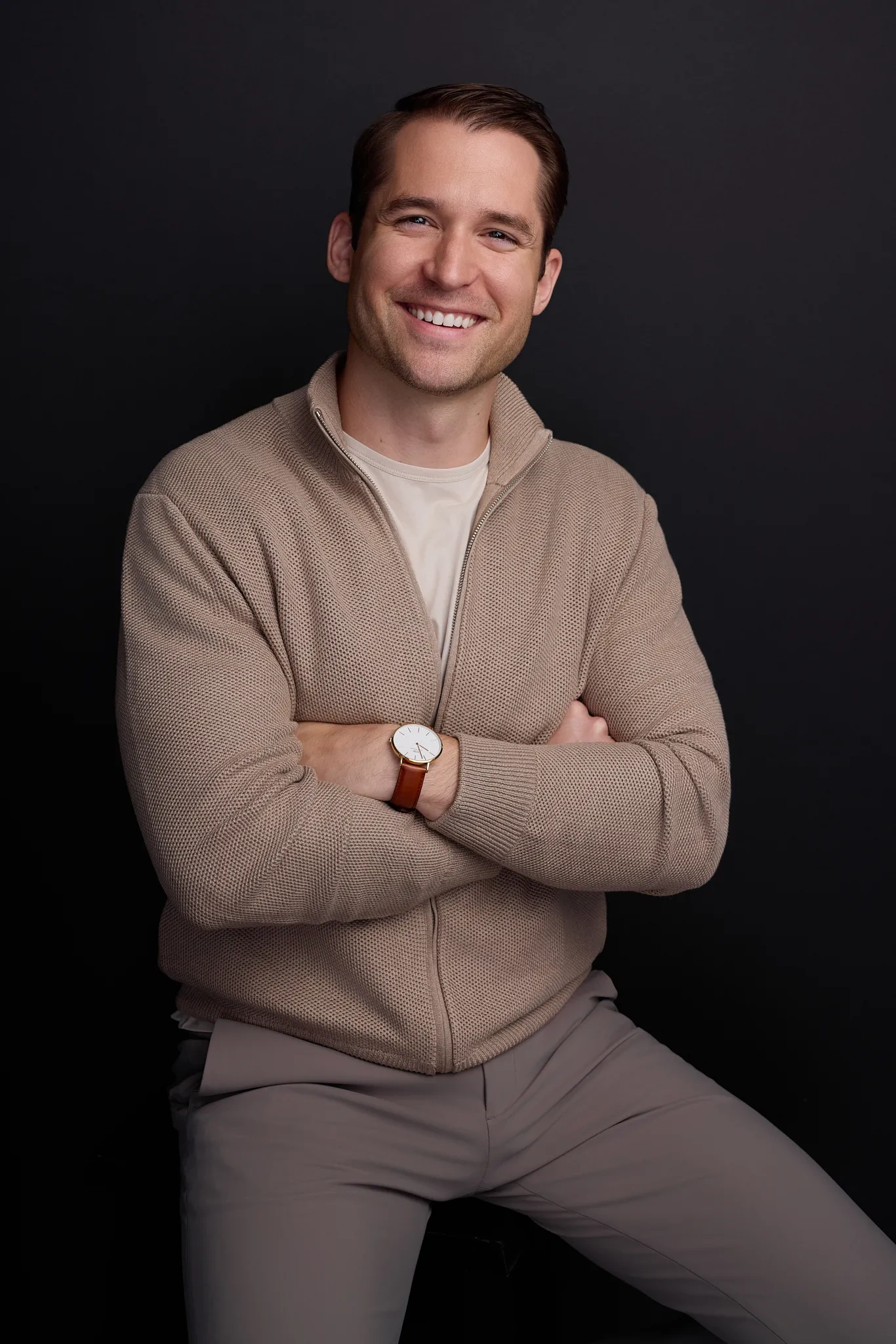 A young white man wearing a tan pullover, a beige T-shirt, and light gray slacks sits on a stool against a dark gray background.