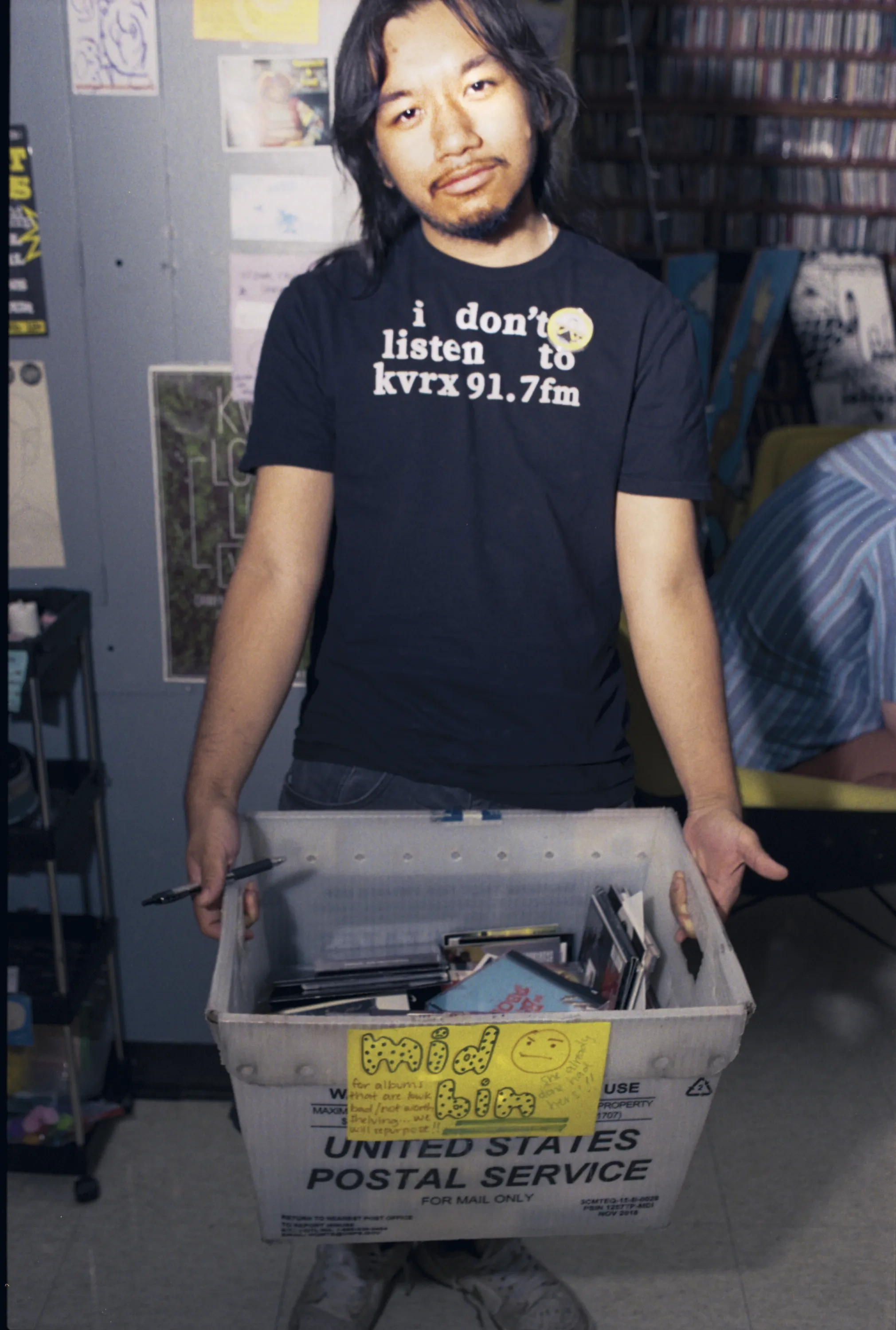 A young man with long hair and a goatee in a black shirt that reads "i don't listen to kvrx 91.7fm" holds a plastic crate of CDs labeled "mid bin."