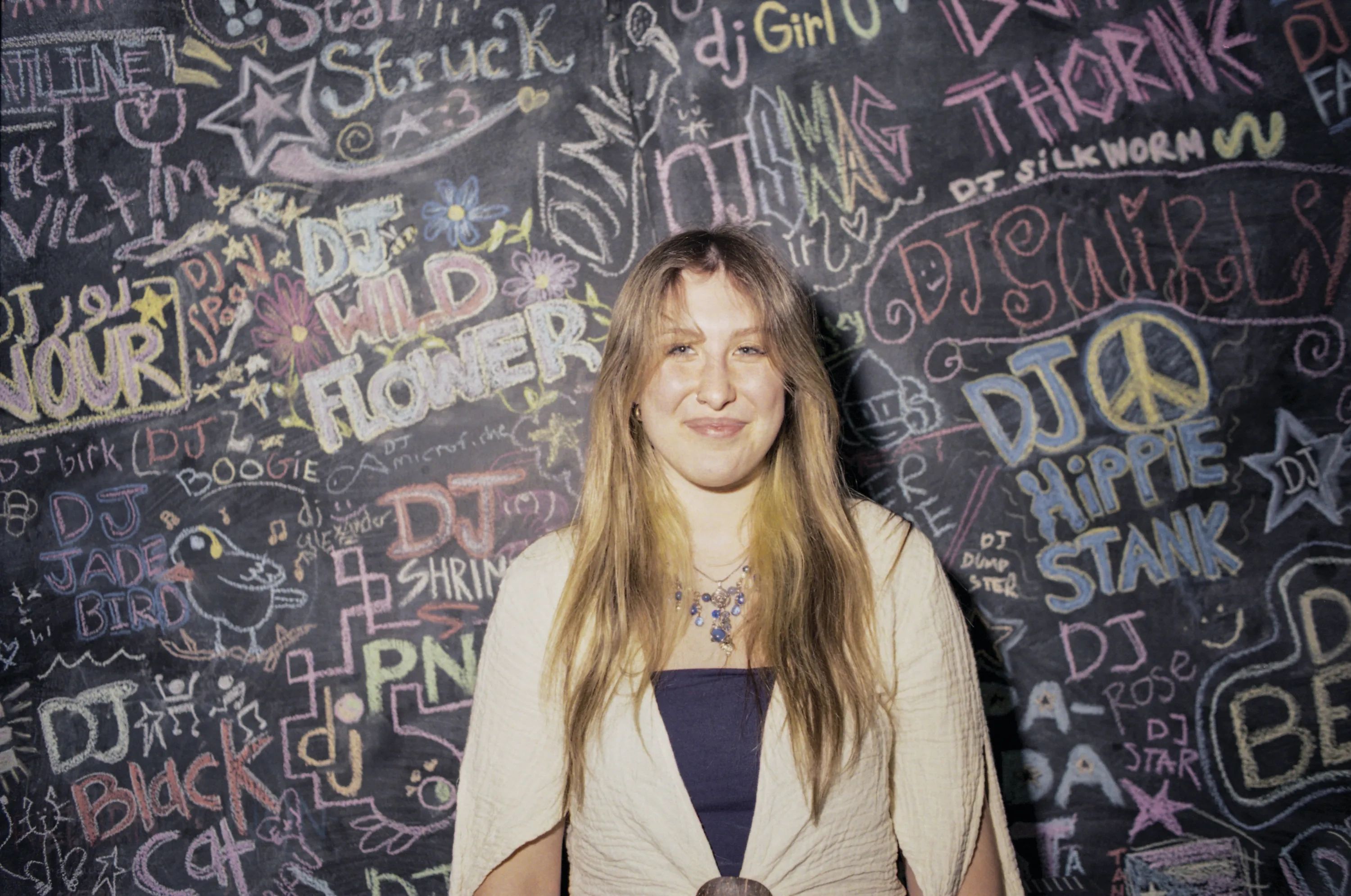 A young woman with blonde hair, wearing a cream cardigan and a funky jeweled necklace, poses against a chalk wall full of graffiti. 