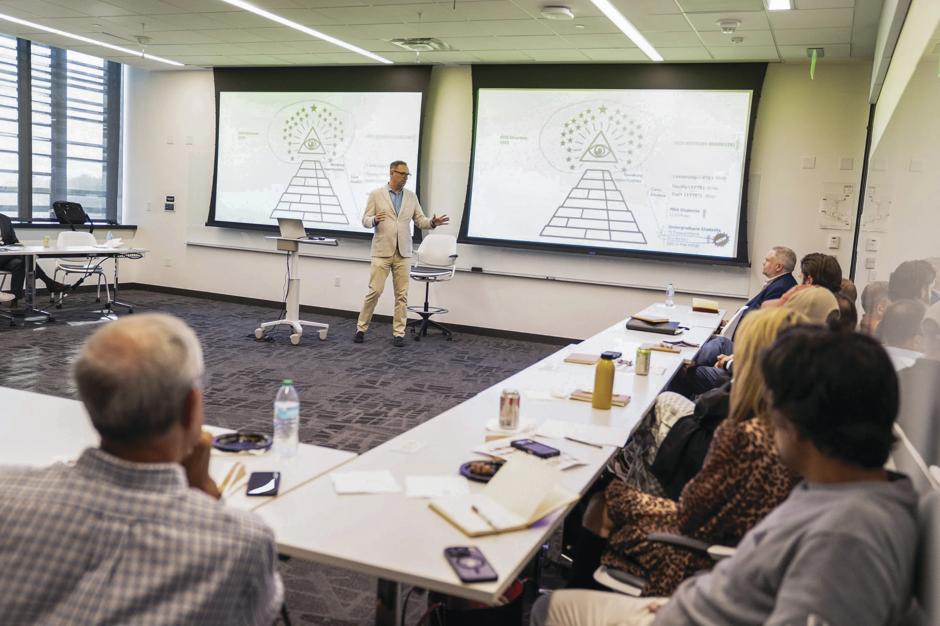 A man in a beige suit presents in front of two projector screens, to an audience of onlookers behind folding tables arranged in an L shape.