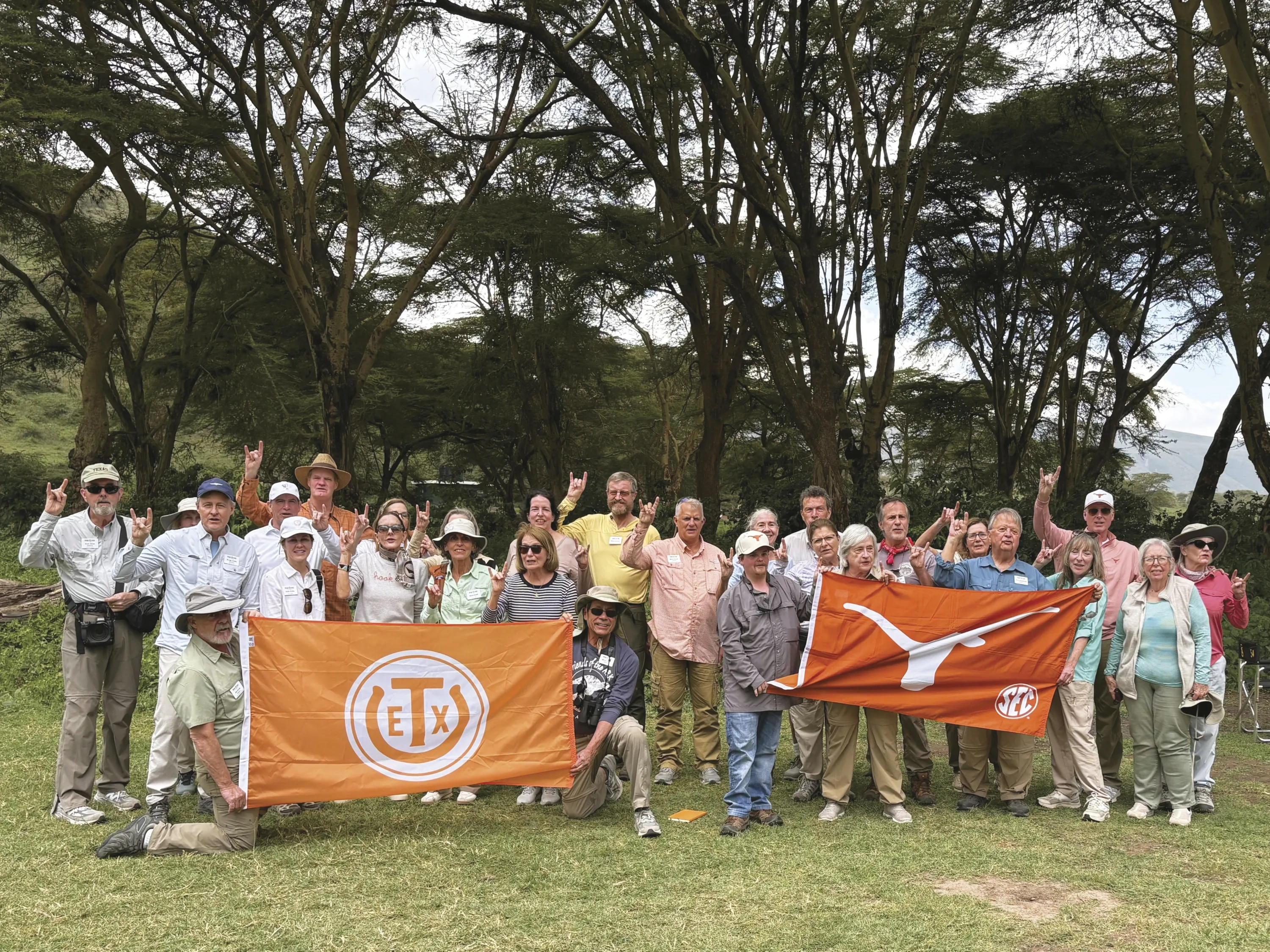 A group of elderly travelers pose in front of a wooded background, holding flags with the Texas Exes logo and Longhorn, respectively.