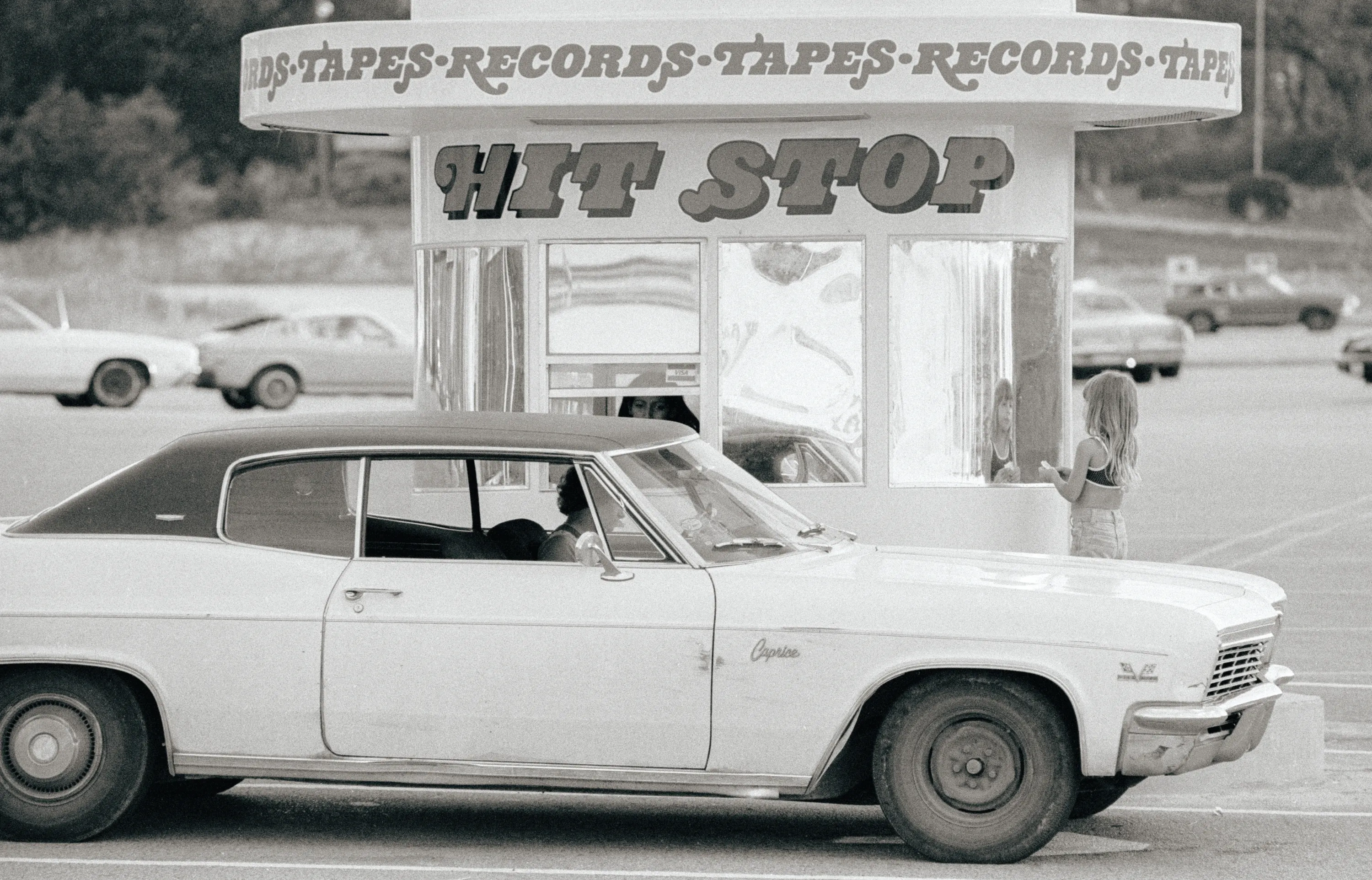 A black and white image of an old car pulled up in front of a drive-through record store with a sign that says HIT STOP in a groovy font.
