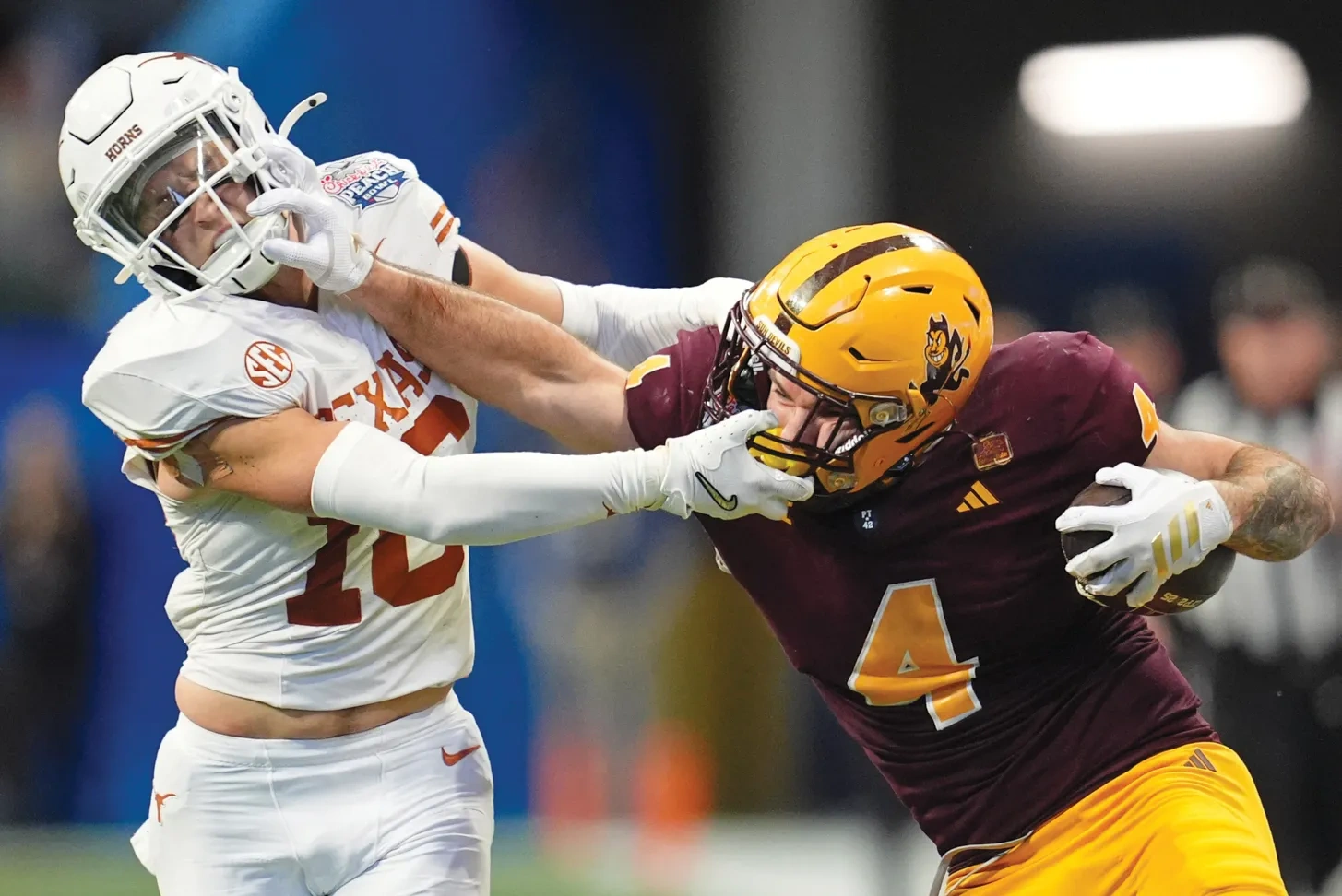 An Arizona State football player in maroon and a Longhorn football player in white have hold of each other's face masks.