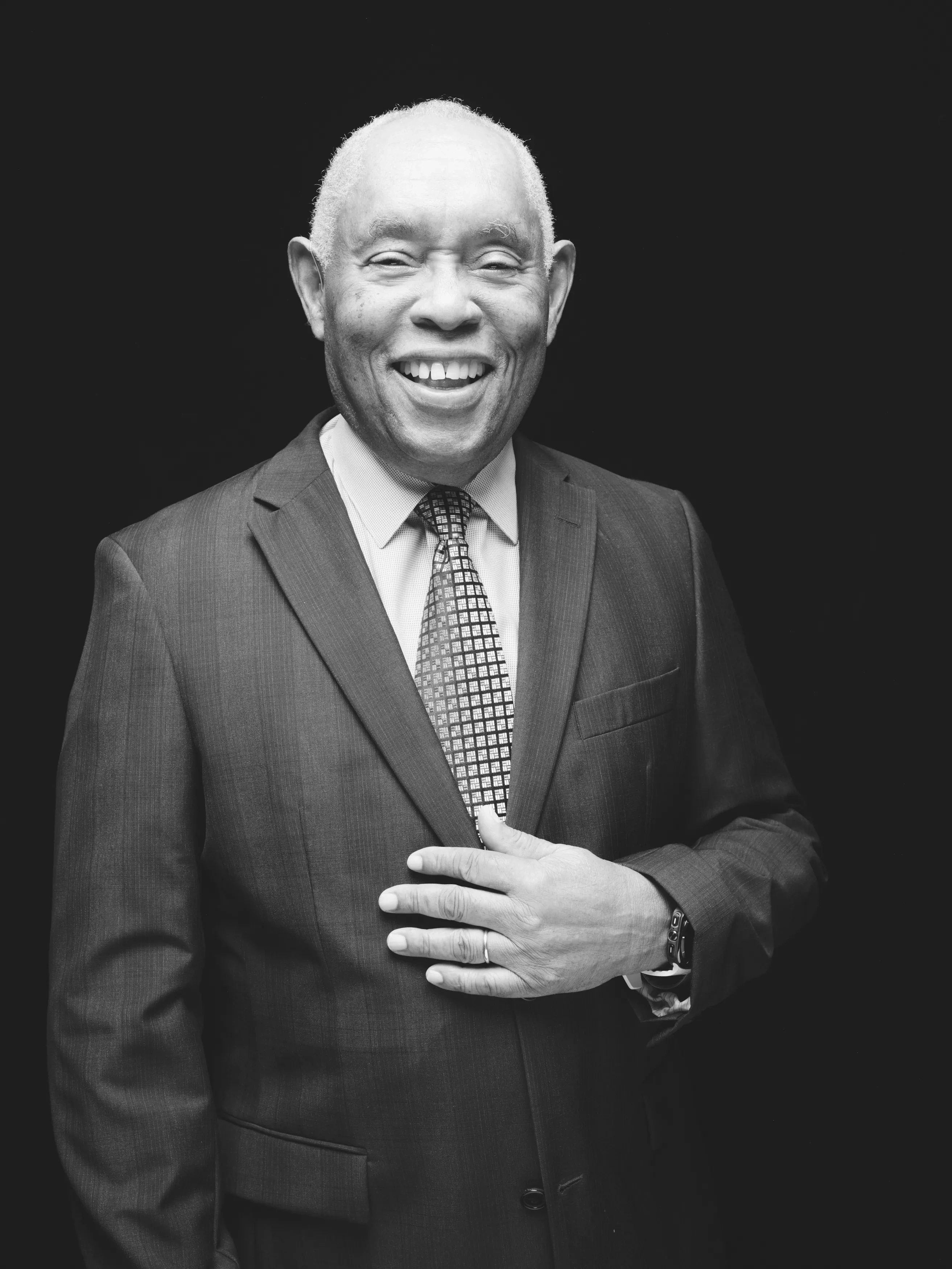 An old Black man stands in a suit and tie with one hand at his midsection, smiling against a black backdrop.