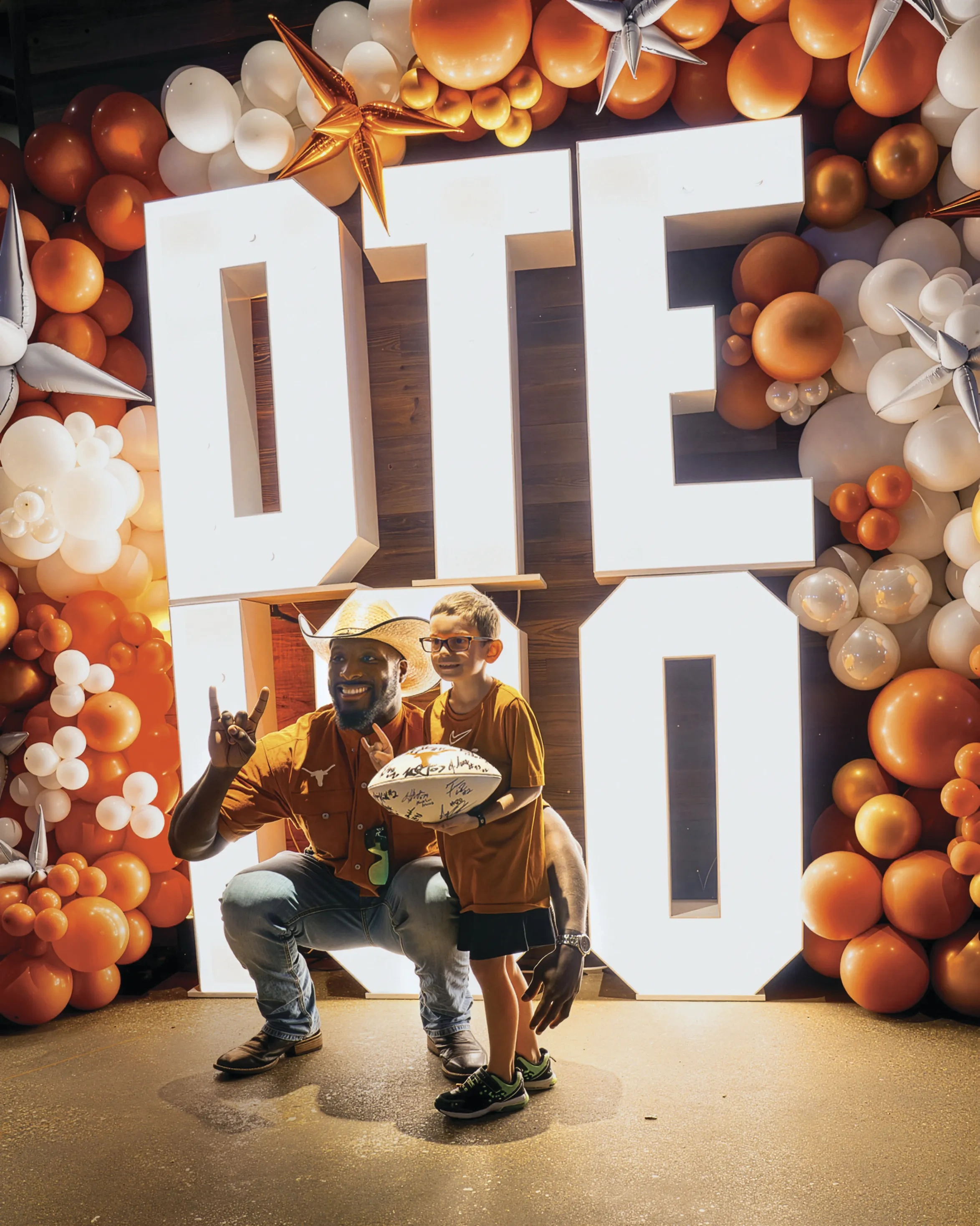 A man in a cowboy hat poses with a child holding a white, signed football. The pair is in front of big, light-box letters that read "DTE 100," surrounded by a burnt-orange and white balloon arch.