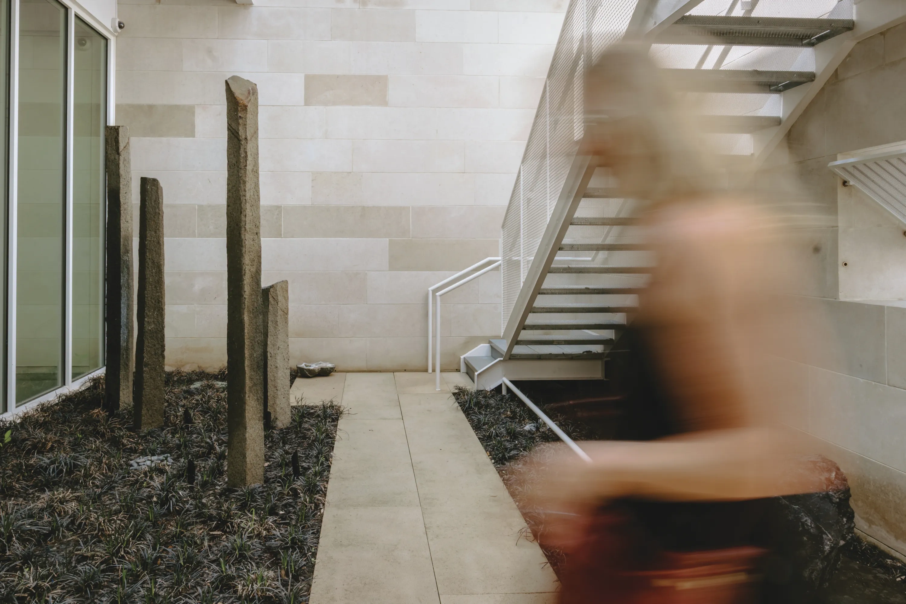 A blurry human figure crosses from the right in a zero-scaped courtyard with vertical stone sculptures.