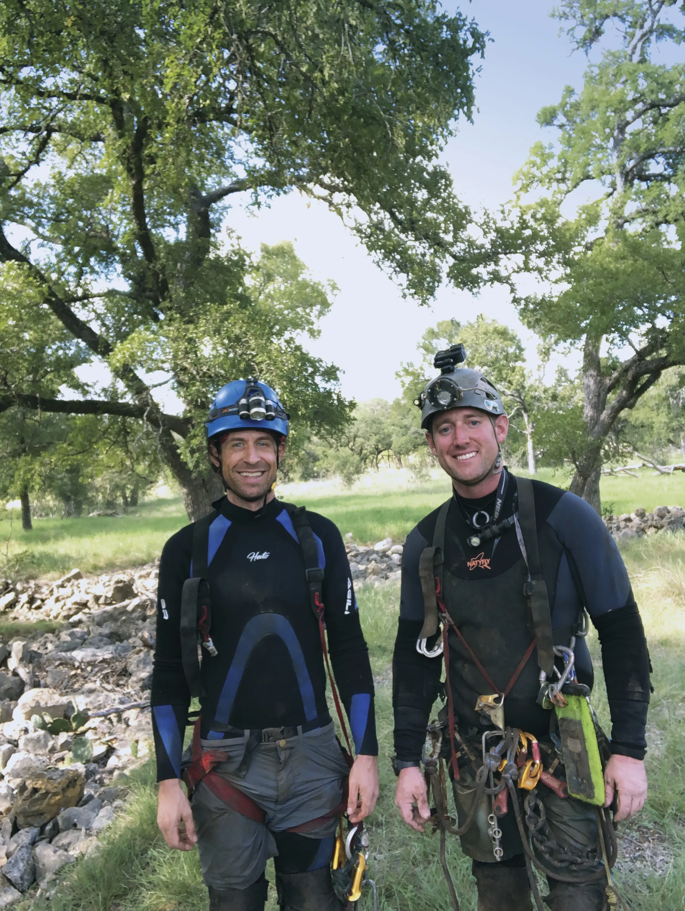 Two men stand in nature in cave-diving gear.