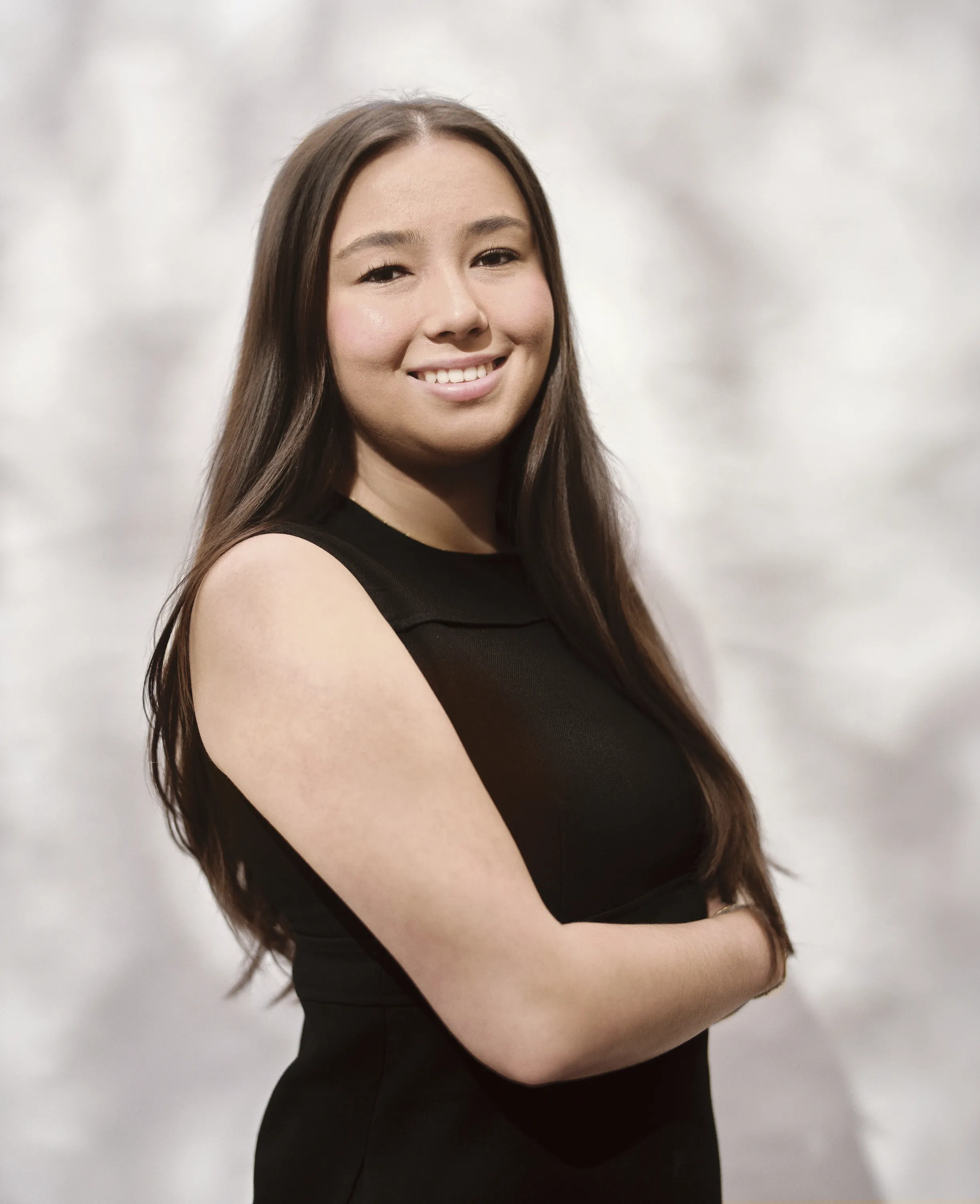 A girl with long dark brown hair in a black dress poses against a dabbled white background.