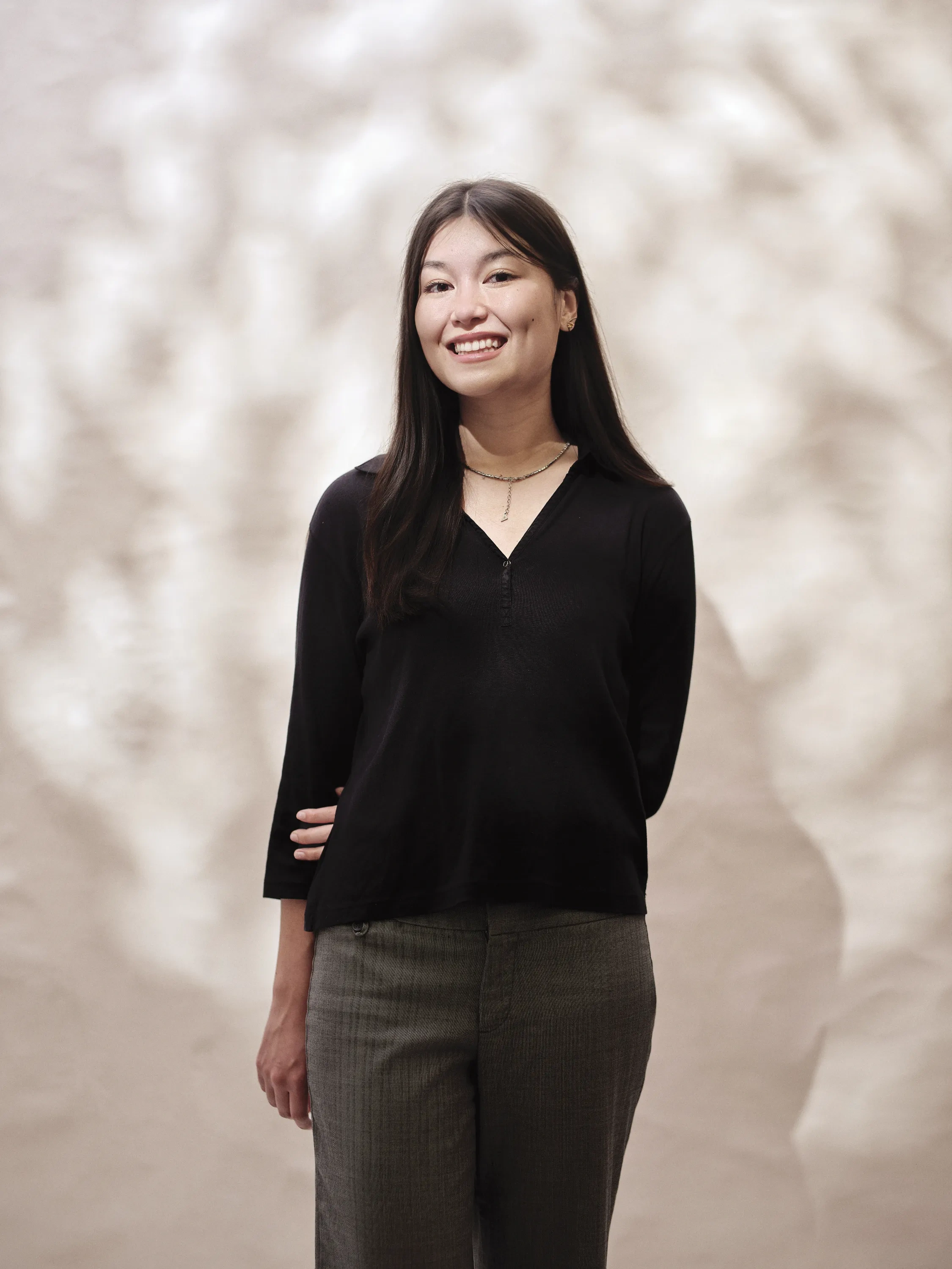 A woman with straight, dark hair, wearing a black blouse and gray slacks, stands in front of a dappled, white backdrop.