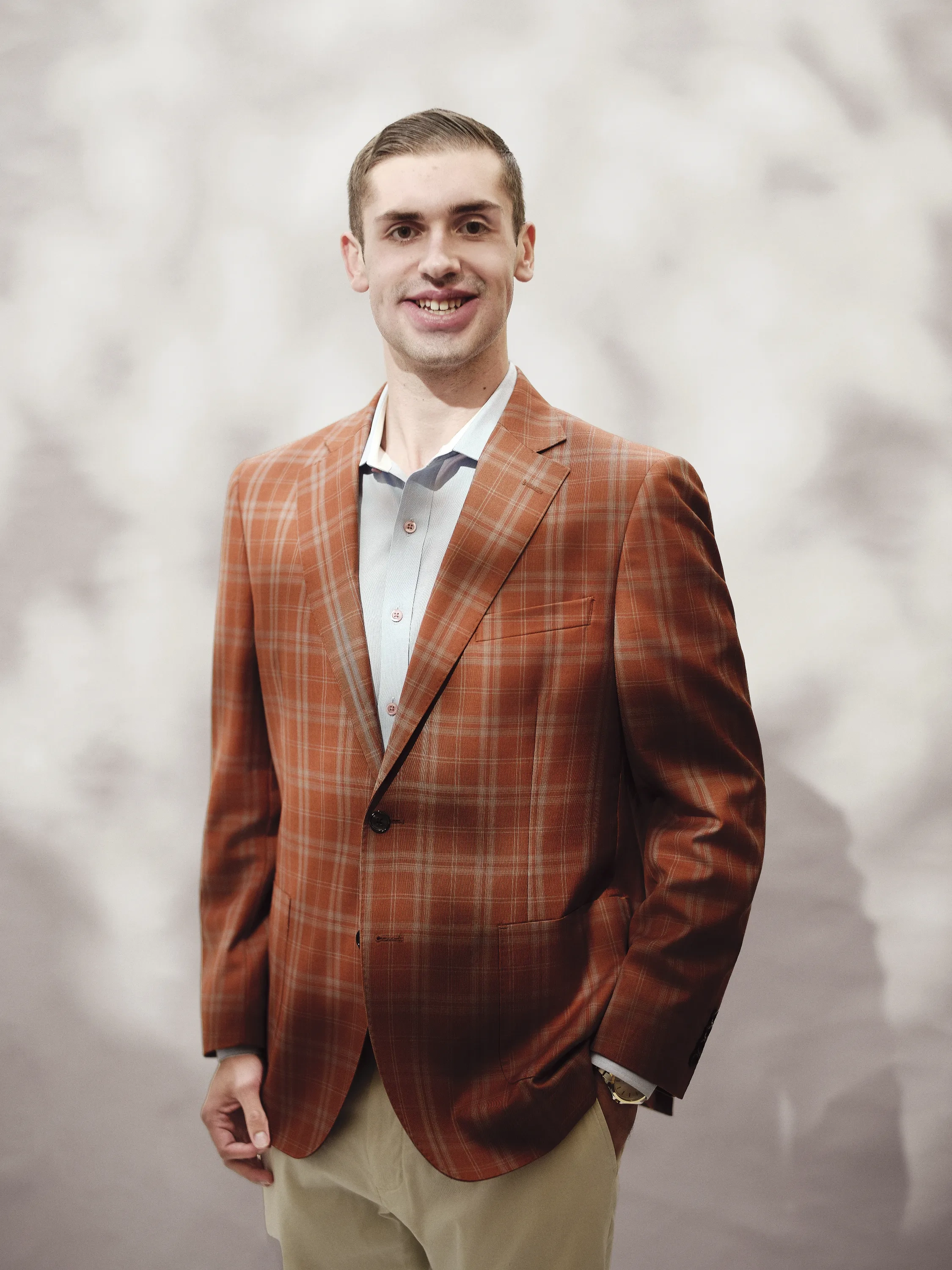 A young man in a plaid burnt-orange blazer poses against a dappled, white background.