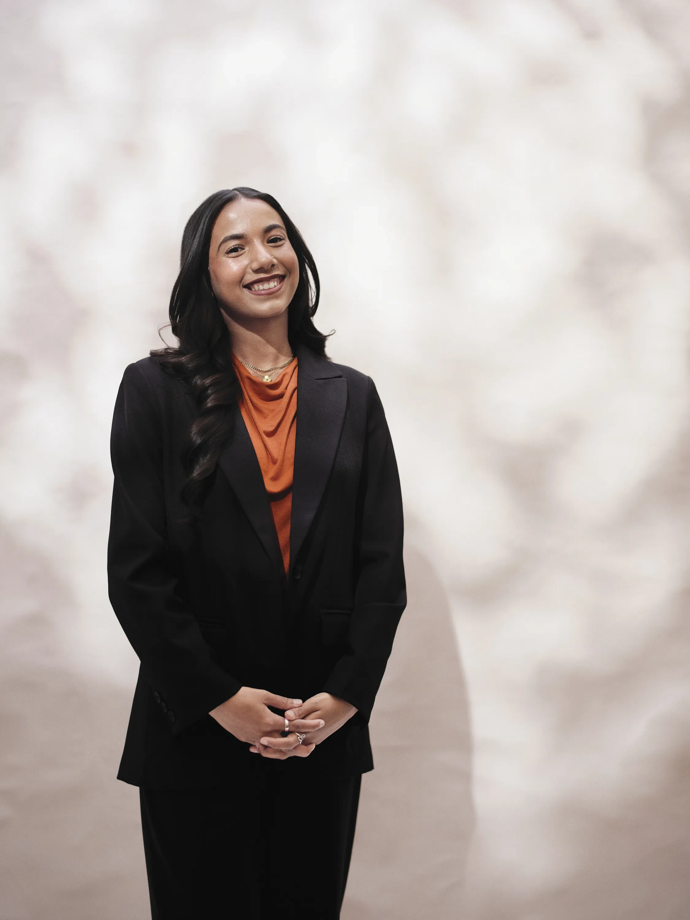 A girl stands in a burnt-orange, cowl-necked blouse under a black blazer poses with her hands clasped in front of her and a smile, in front of a dappled, white background.