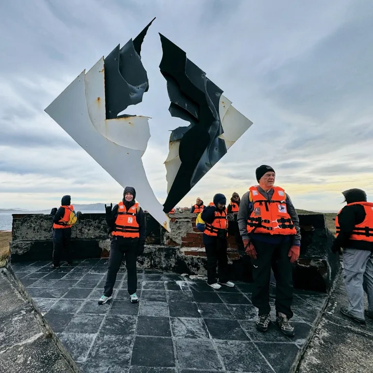 A woman in an orange vest stands in front of a sculpture on Cape Horn.