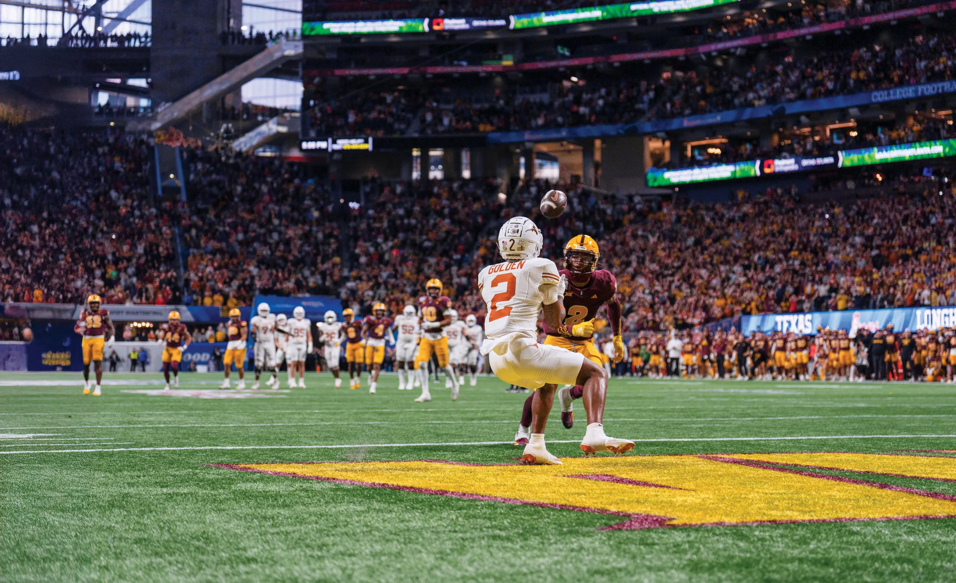 A Longhorn football player in white catches a football ahead of a football player in maroon.