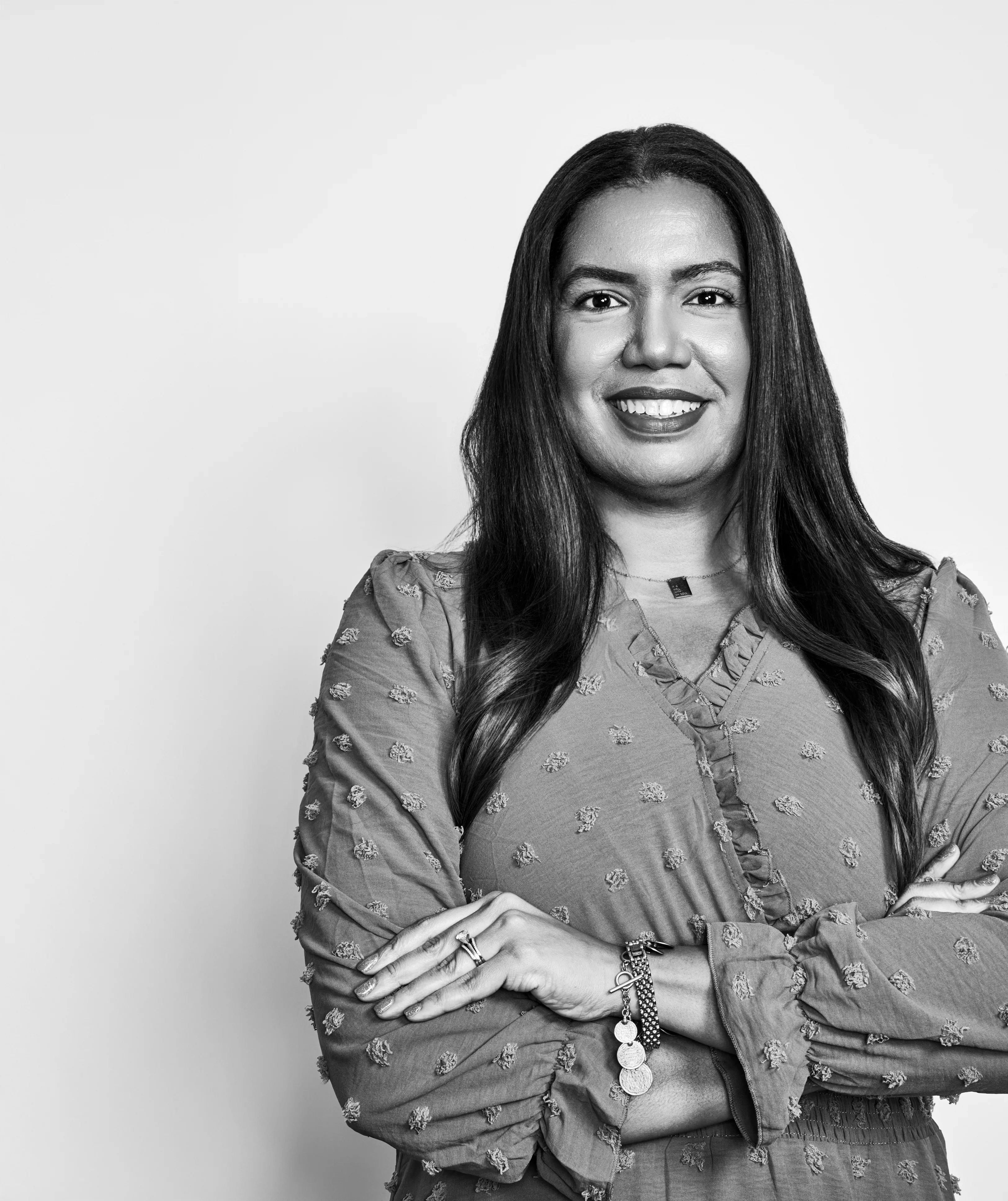 A brown-skinned woman with her hair down poses with her hands folded across her chest.