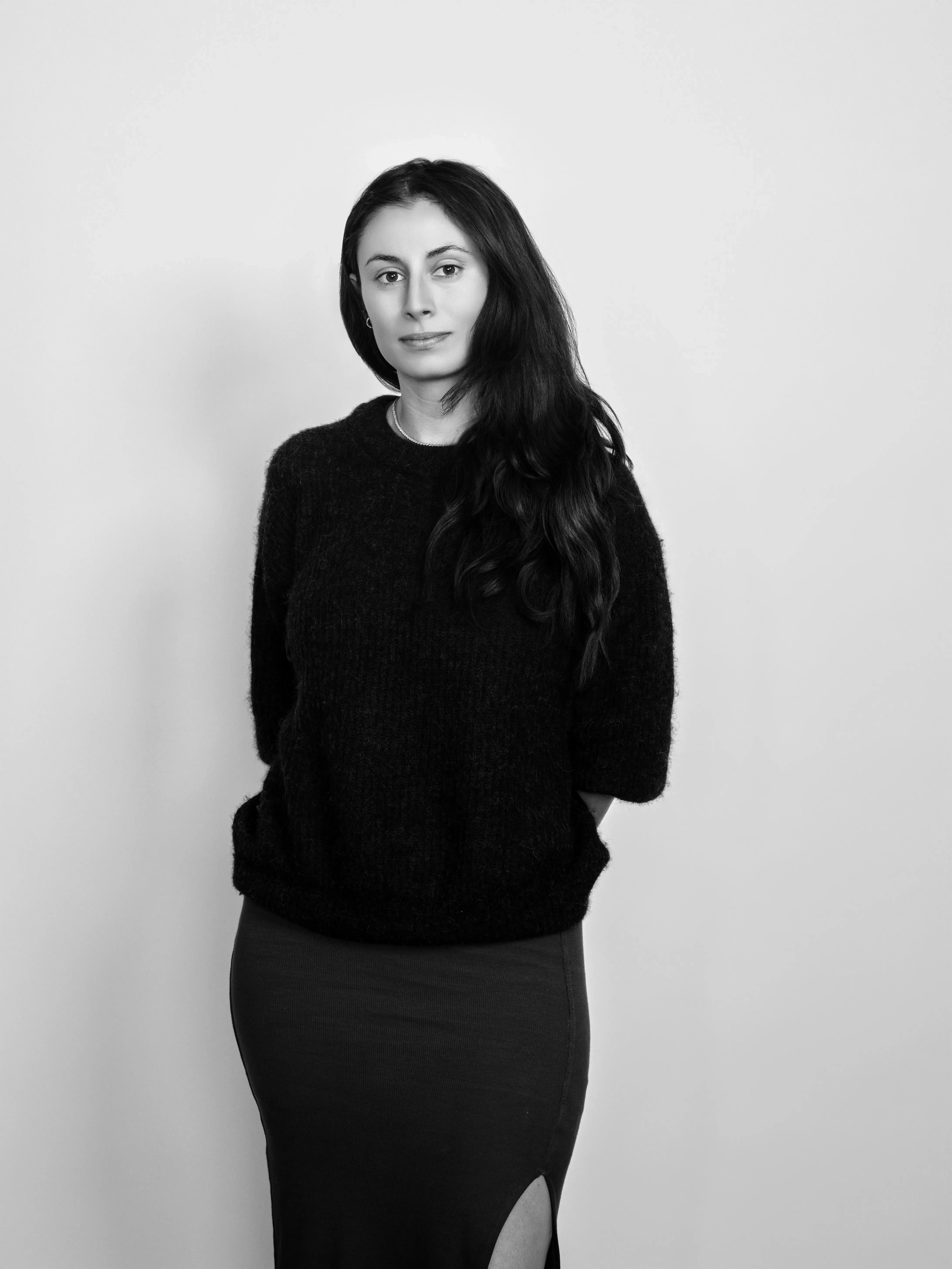 A woman poses with her hands behind her, wearing a sweater over a skirt.