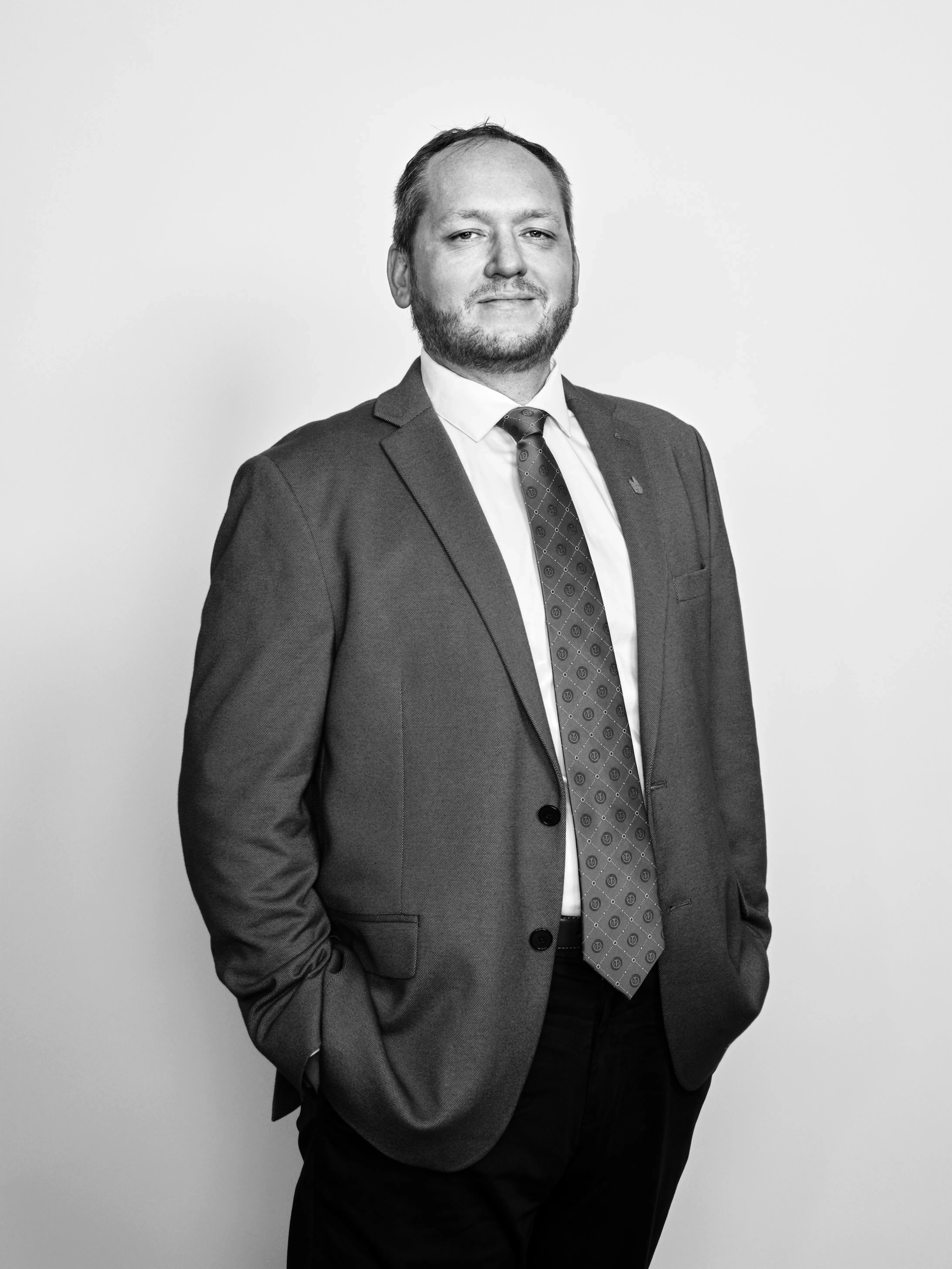 A man with some facial hair poses with his hands in his pants' pockets, wearing a tie and sports coat.
