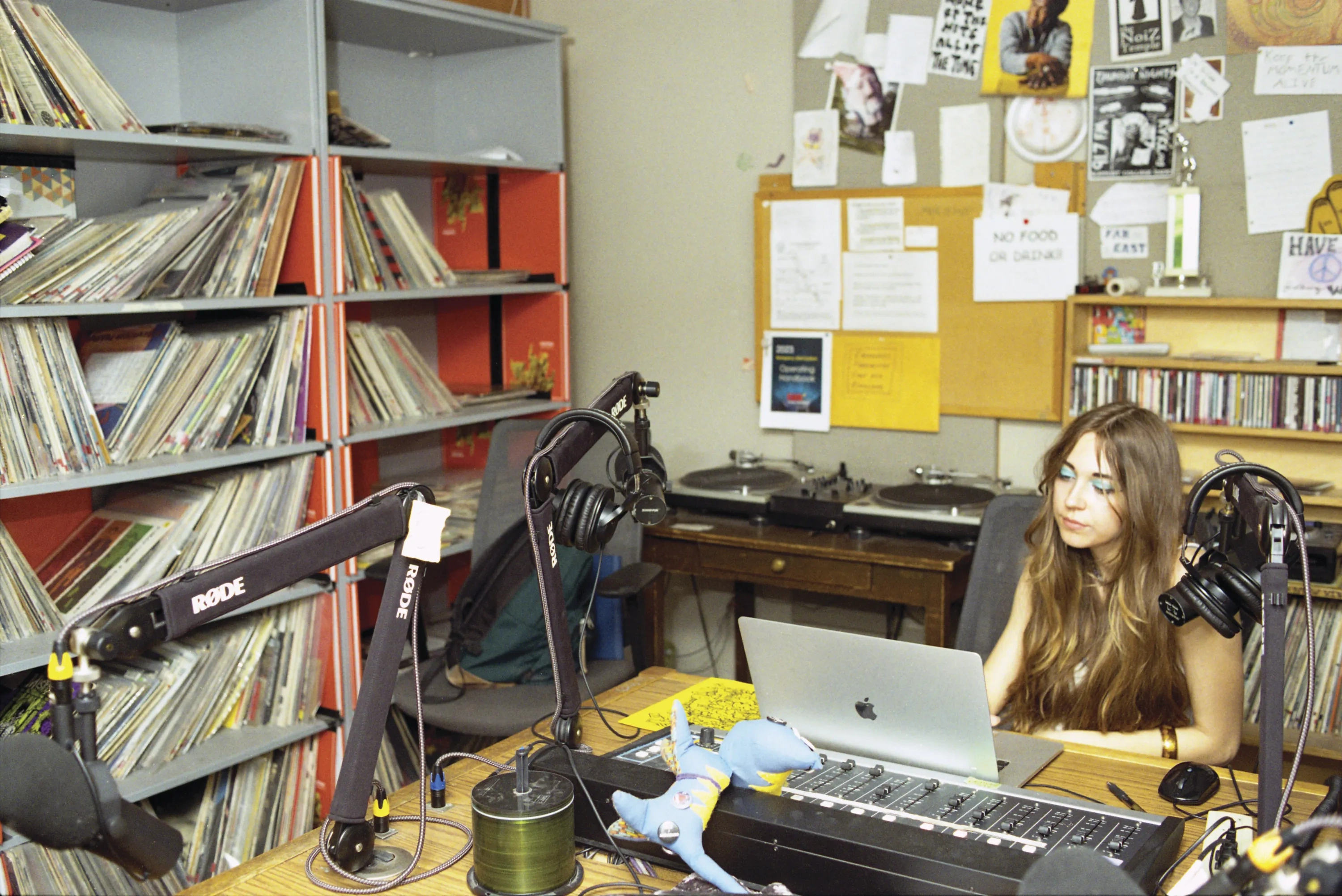 A young woman sits at a radio DJ desk, next to a packed shelf of vinyl records.