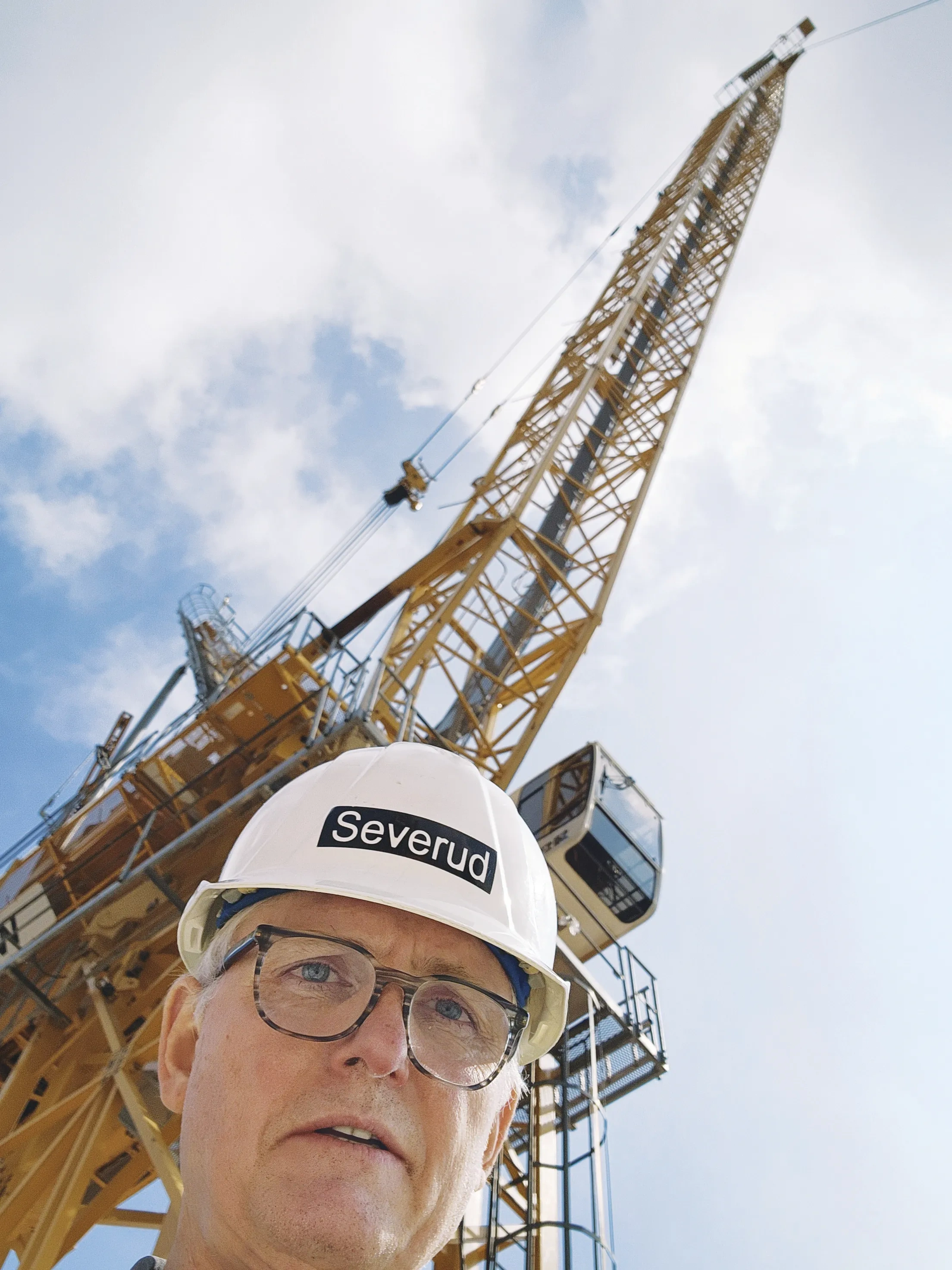 A man in a white hard hat that reads "Severud" takes a selfie in front of a large yellow crane against a cloudy blue sky.